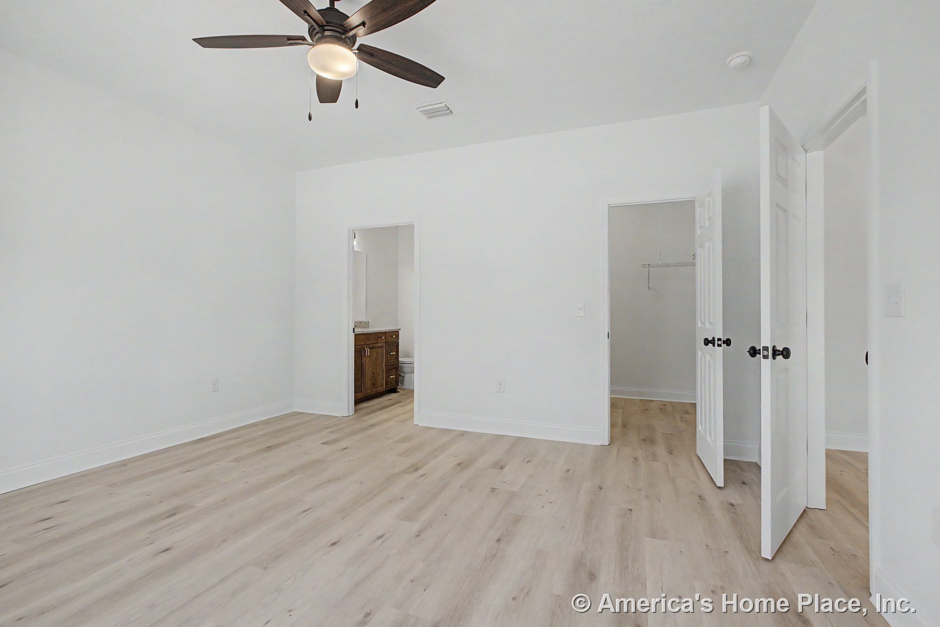 Bedroom with light wood plank flooring, white painted walls, ceiling fan with integrated lighting, direct access to bathroom featuring granite vanity, walk-in closet with shelving