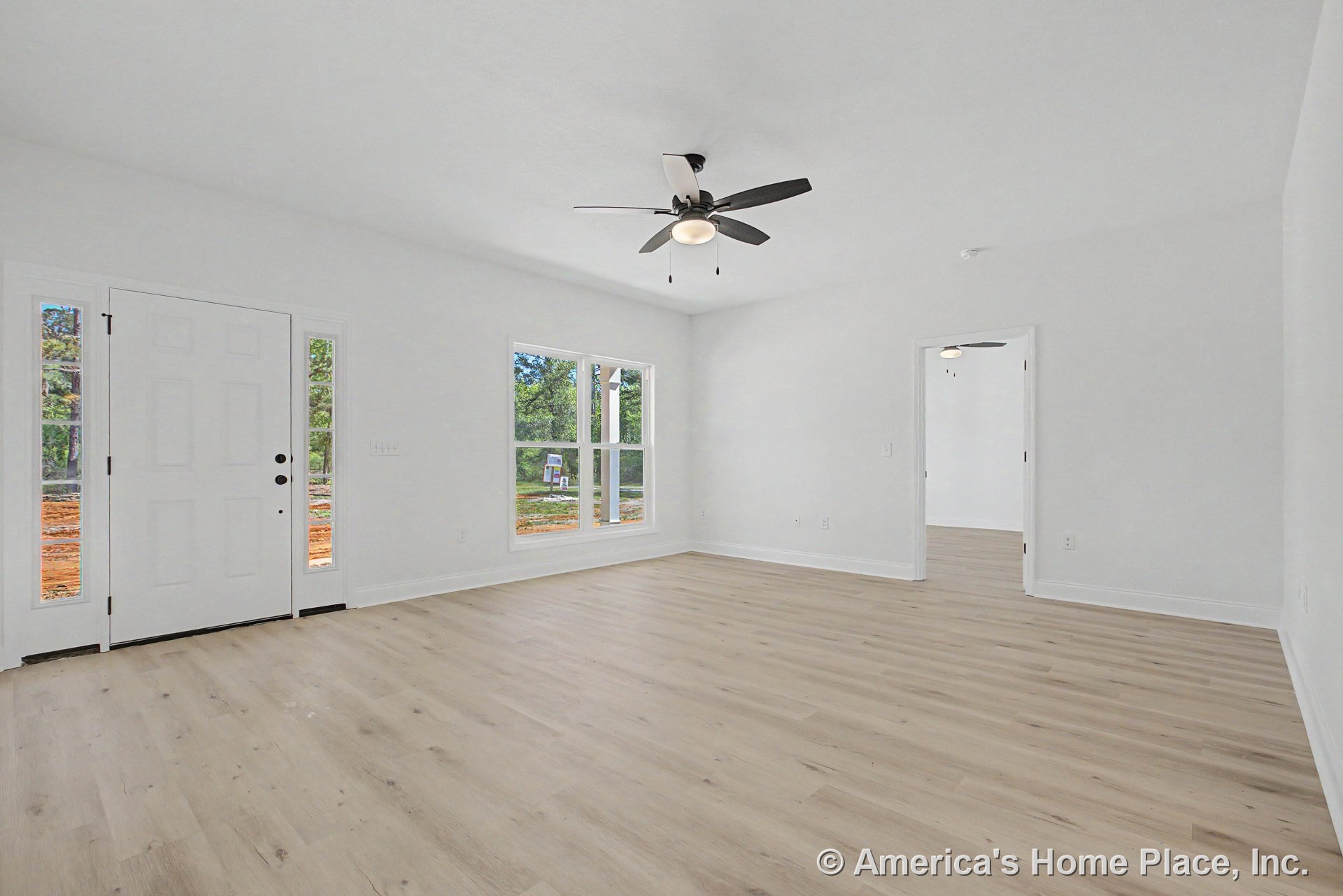 Light wood flooring and white baseboards in a spacious living room with large central window, double front door with sidelights, smooth white ceiling, modern ceiling fan with