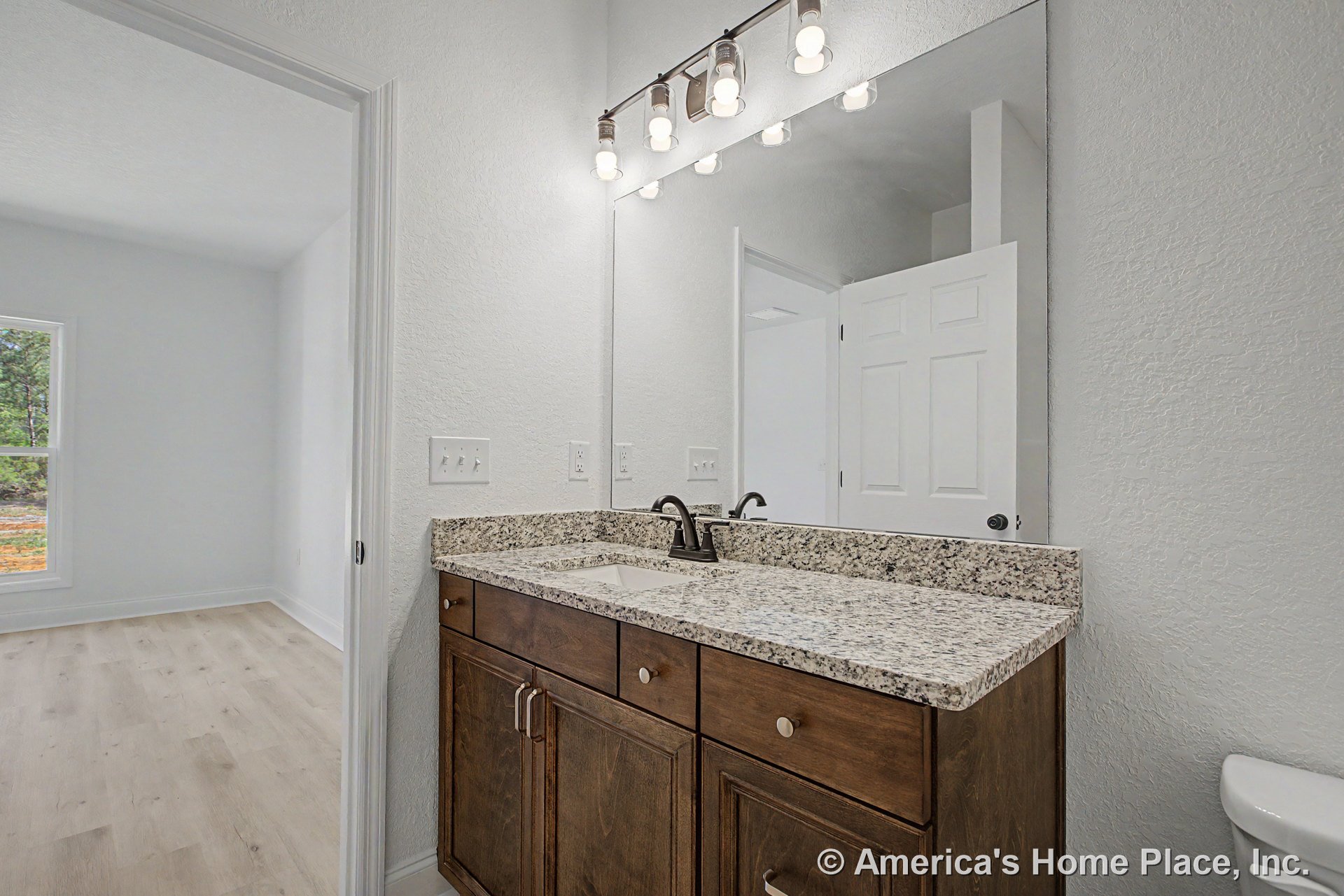 Granite vanity countertop with dark wood cabinetry, rectangular undermount sink, large wall-mounted mirror, modern vanity light fixture, white textured walls, light flooring, and
