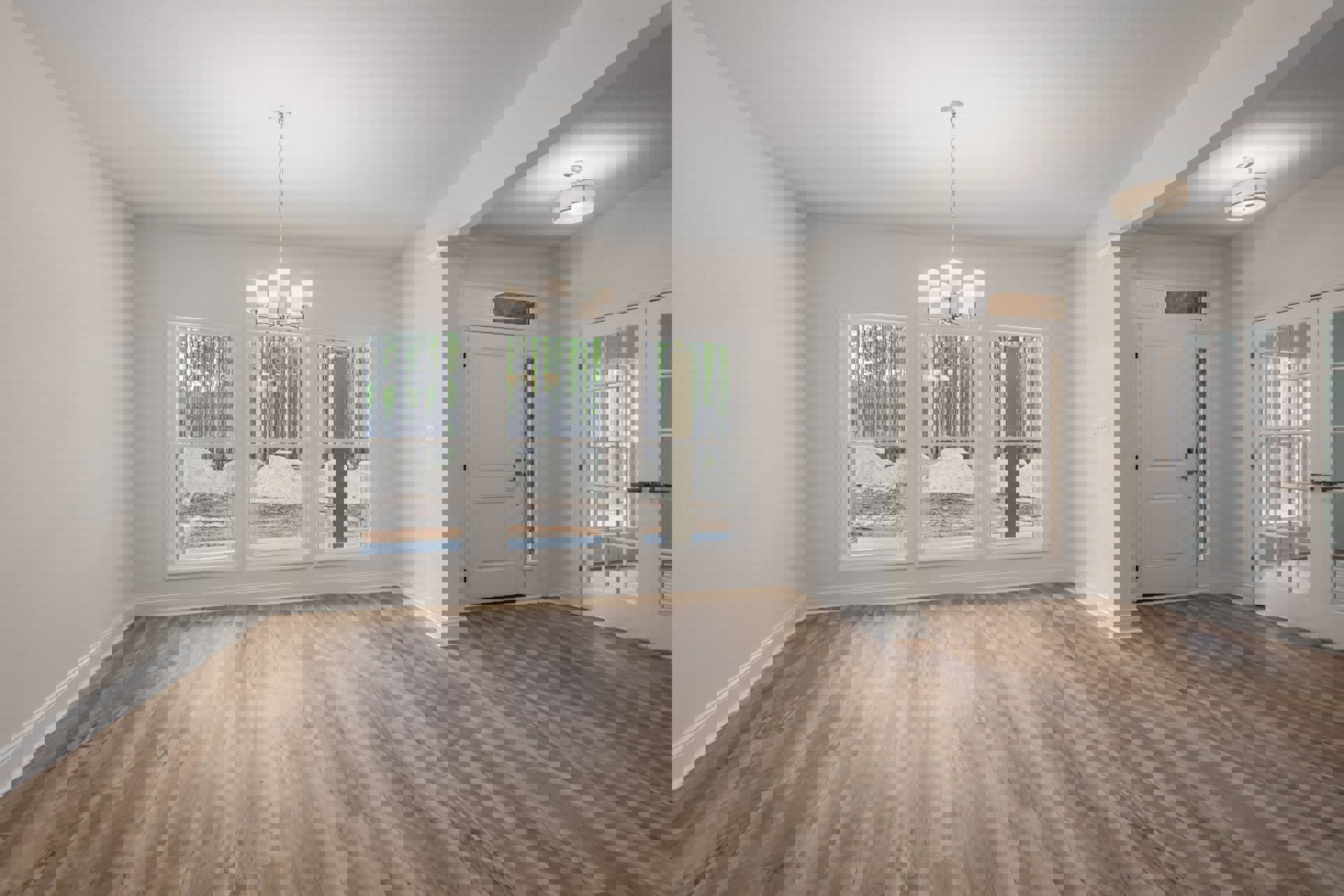 Wood flooring in a spacious room featuring a modern chandelier, white door with metal doorknob, large windows overlooking a forest, and smooth plaster walls.