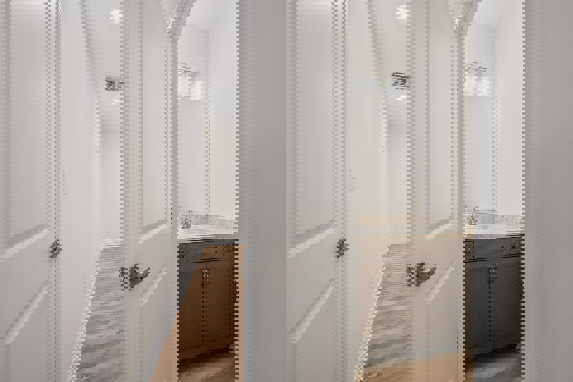 Bathroom with wood flooring, white sink set in a brown cabinet with marble countertop, white ceiling light, white walls, and a white door; floor vent visible near the door.