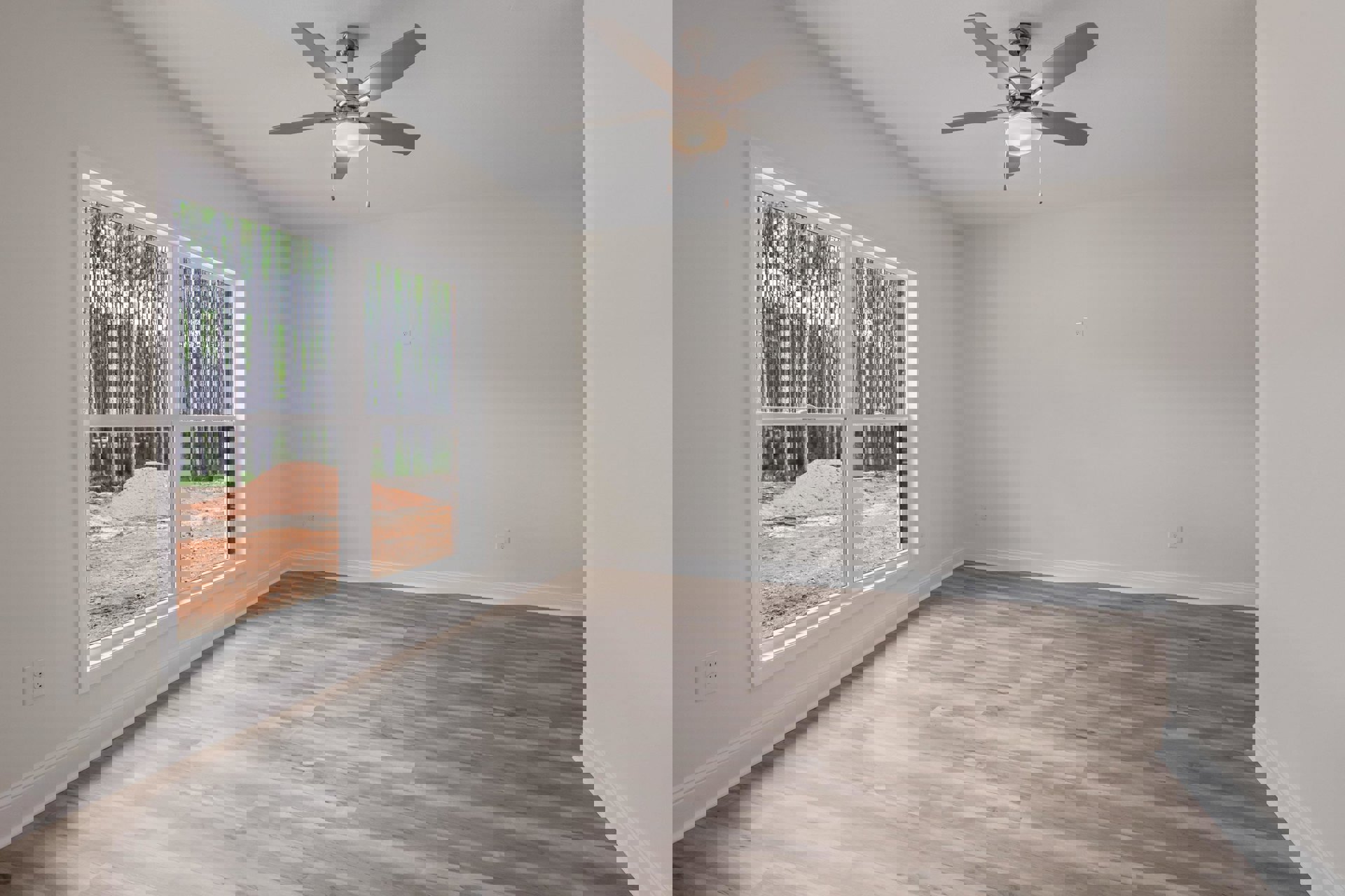 Ceiling fan with light fixture above wood flooring, large window overlooking trees and dirt pile, white plaster walls and ceiling.