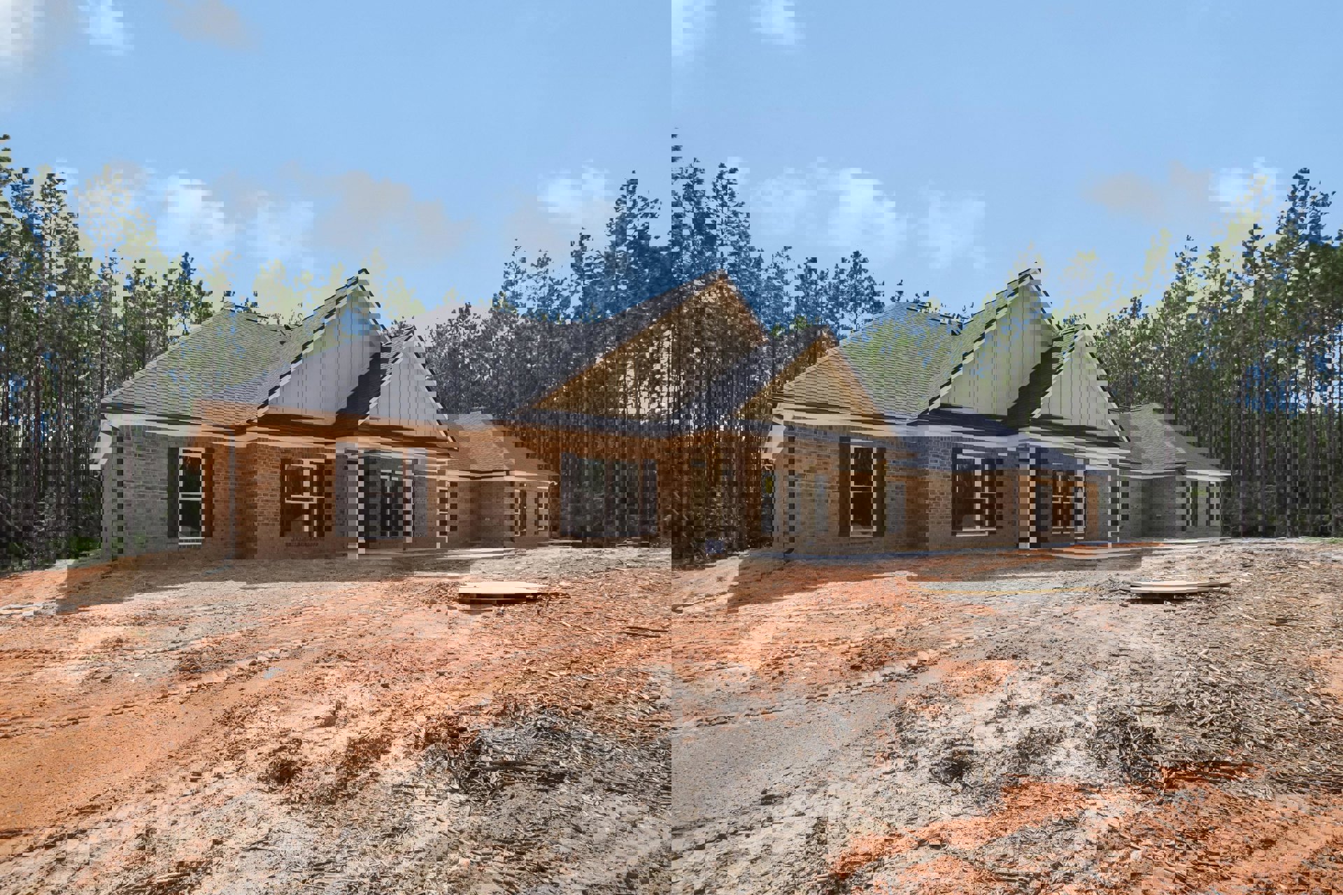 Partially built brick house with white columns, black shuttered windows, and white-framed windows, surrounded by trees and a dirt road with tire tracks