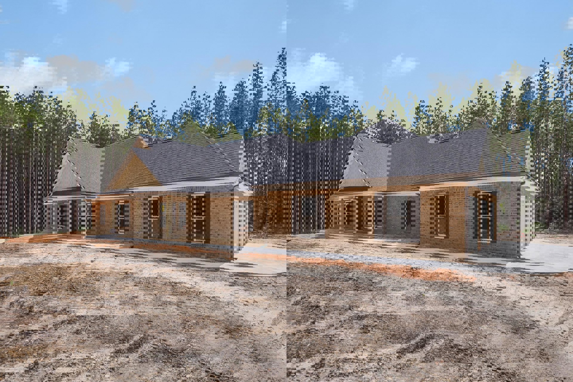 Partially built brick house with white-framed windows, screened door, broken glass pane, dirt and debris scattered on bare ground, surrounded by tall trees under a blue sky.