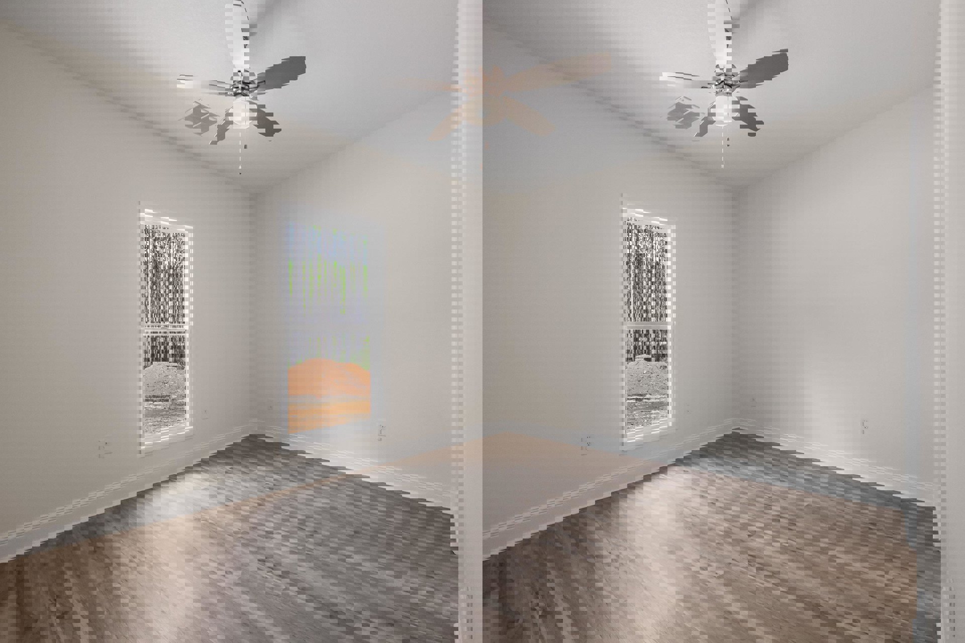 Wood floor room with white plaster walls, ceiling fan with light, large window overlooking dirt hill and trees.