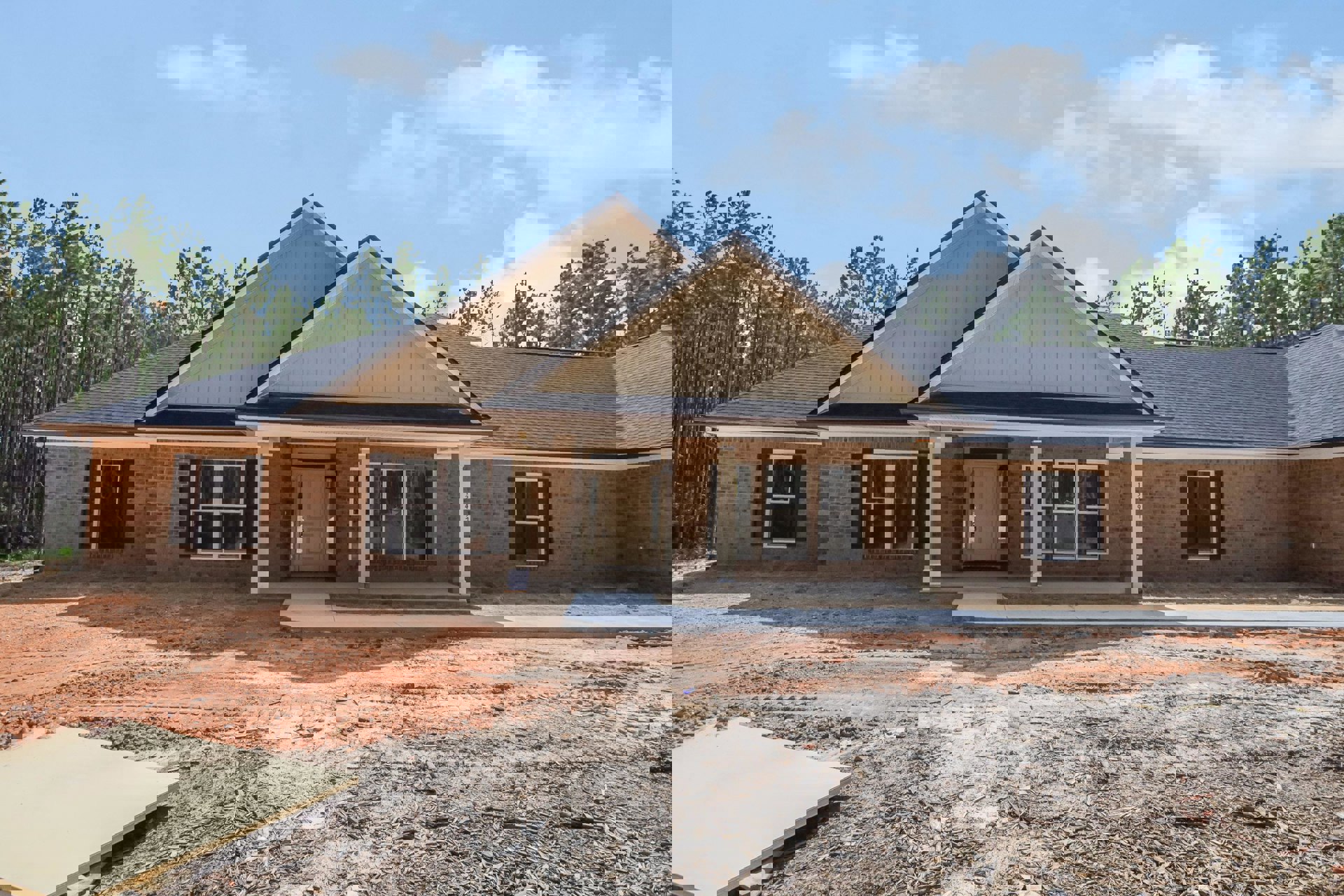 Partially built house with exposed framing, white door, white-framed window, dirt and wood chips covering the ground, surrounded by trees under a cloudy sky