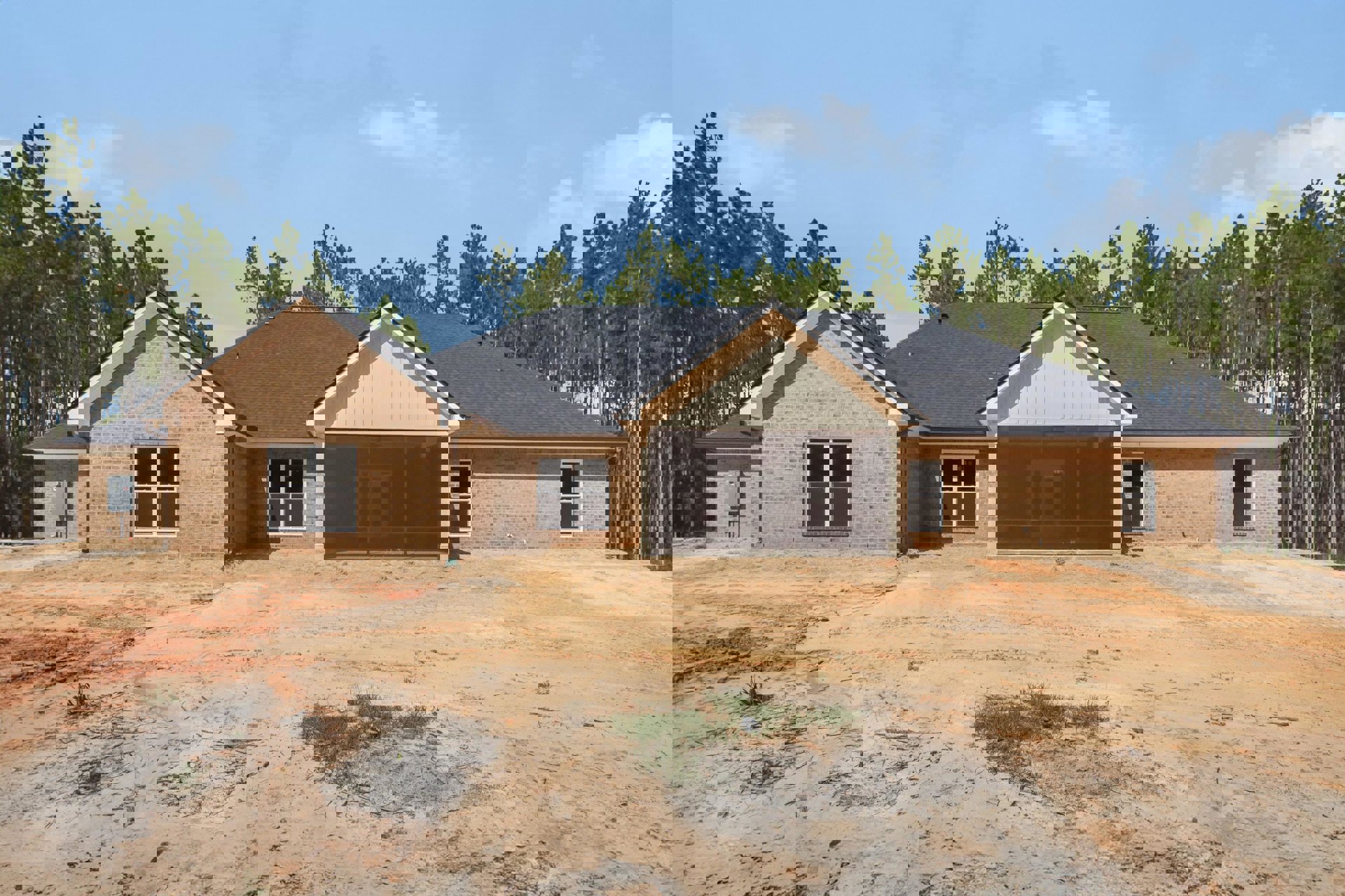 Partially built brick house with white-framed windows, exposed roof structure, dirt yard bordered by grass, and mature trees in the background