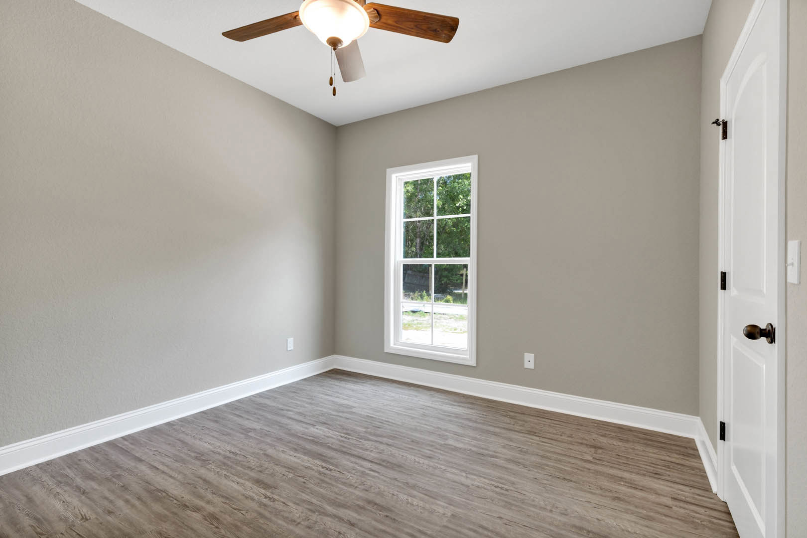 Bedroom with wood flooring, white baseboards, ceiling fan with light fixture, white-framed window, and white door featuring black hinges