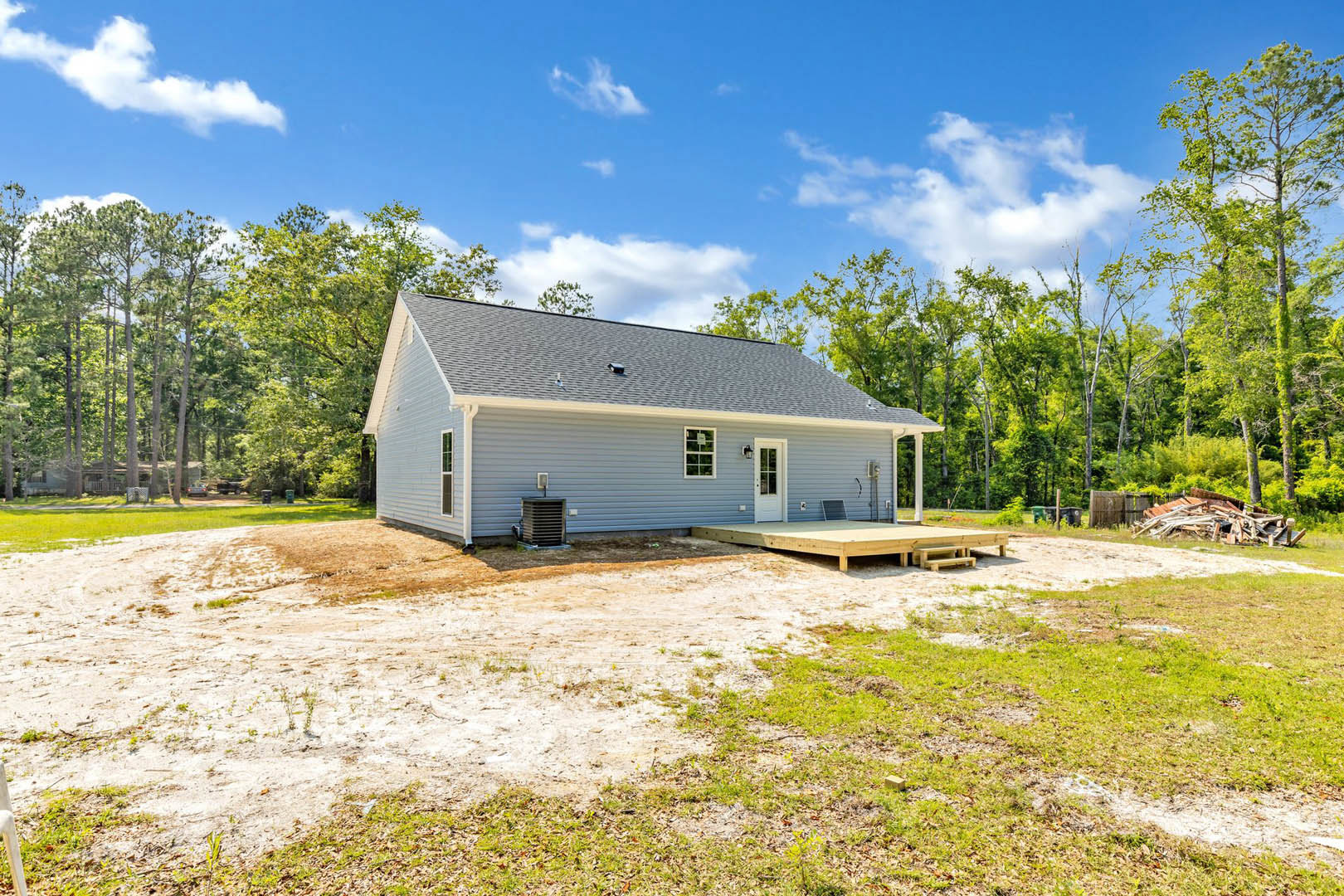 Two-story house with light siding, white front door, covered porch, wooden deck elevated on posts, dirt yard with scattered wood, grassy area, trees in background, cloudy sky