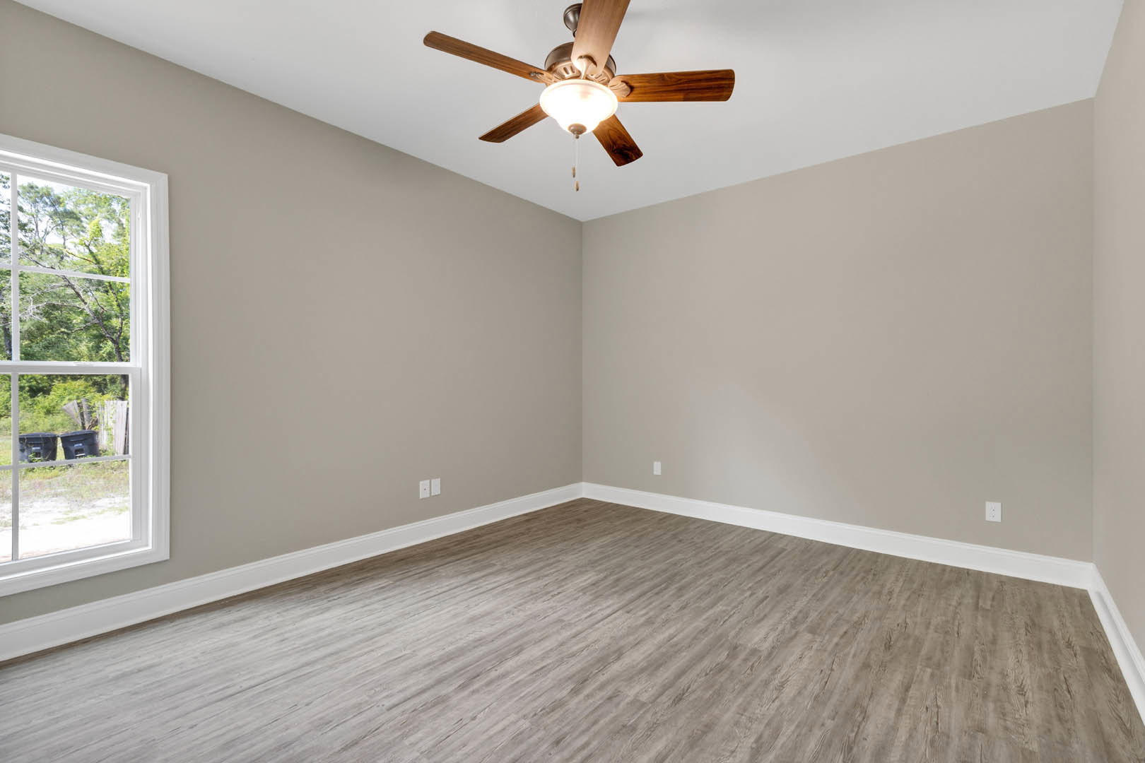 Ceiling fan with light fixture mounted on white plaster ceiling above wood laminate flooring, window with book and basket on sill, white baseboards along wall, black trash can