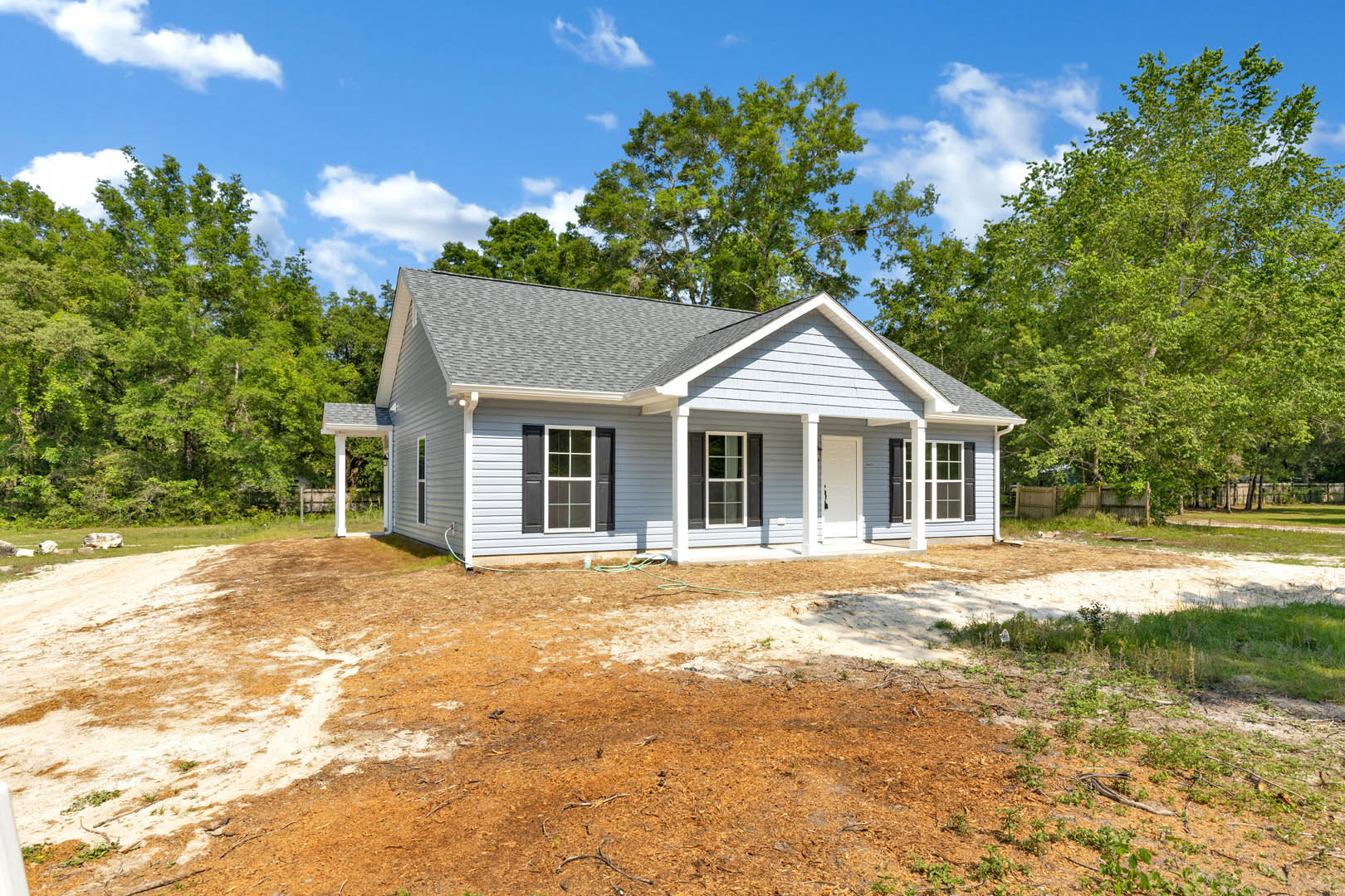 Two-story house with white-trimmed windows, covered front porch, dirt yard in foreground, and mature trees in the background