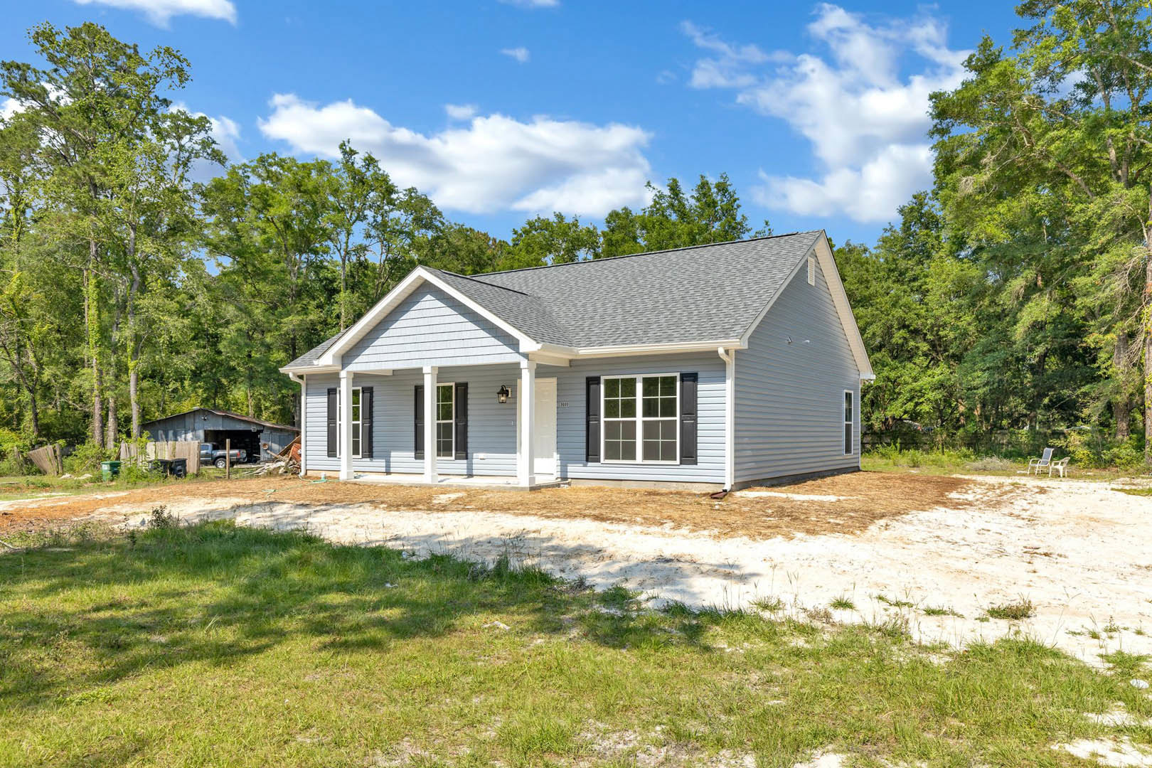 Two-story house with white door and black shuttered window, surrounded by dirt and mature trees, white lawn chair on grassy area, cloudy sky overhead
