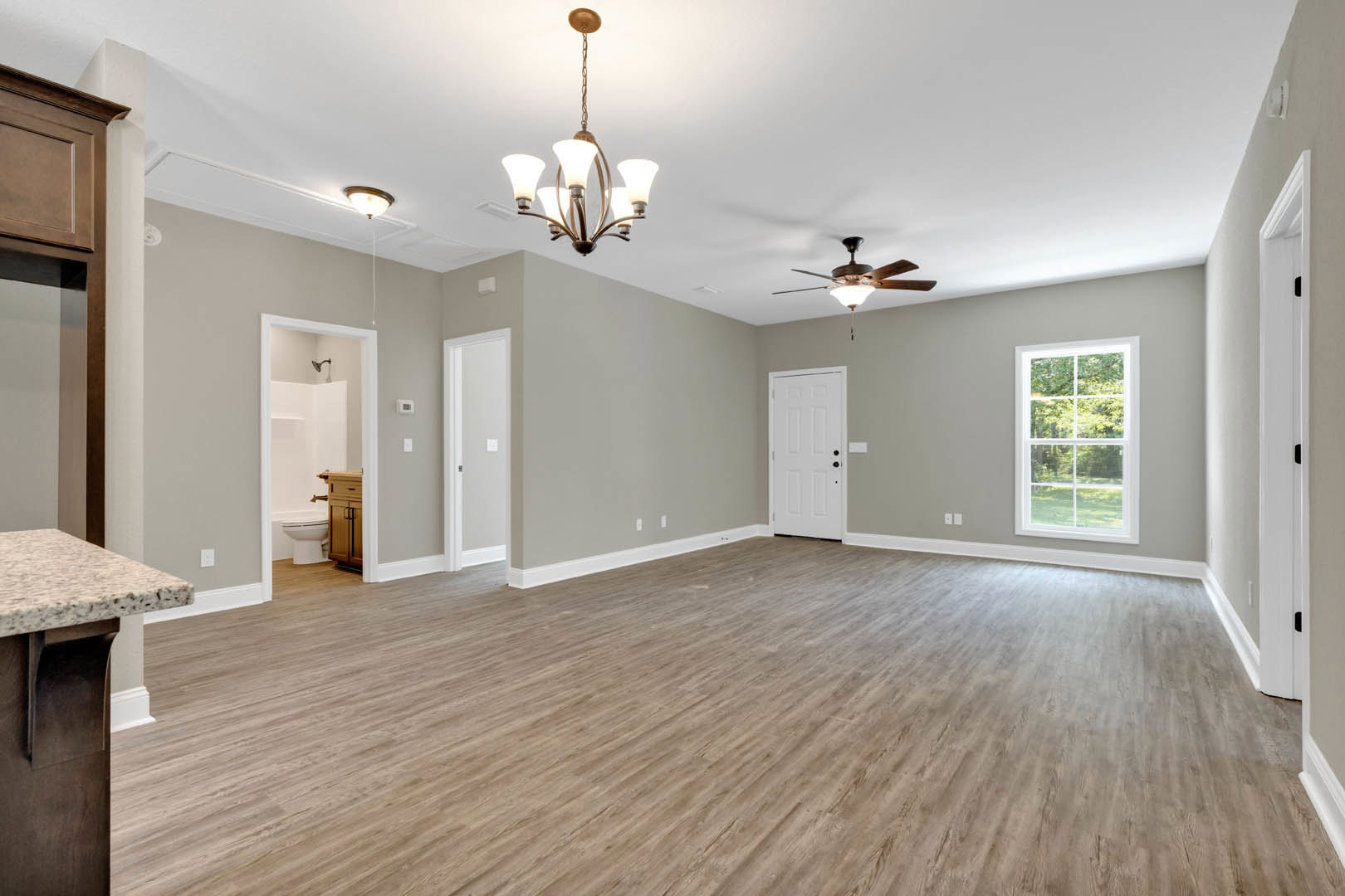Spacious room with hardwood flooring, multi-pane window, white door with black hardware, ceiling fan, and chandelier; close-up of stone countertop visible.
