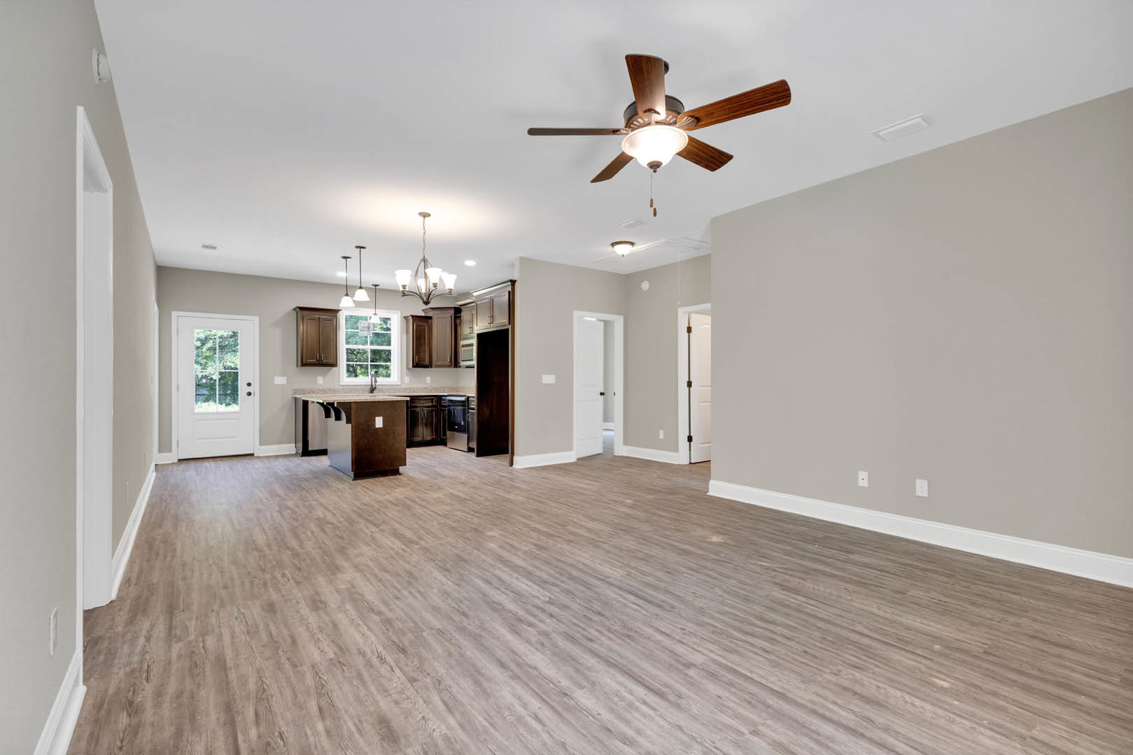 Open-concept room featuring a ceiling fan with light fixture, hardwood flooring, white paneled door with black hardware, and kitchen area with stone countertop