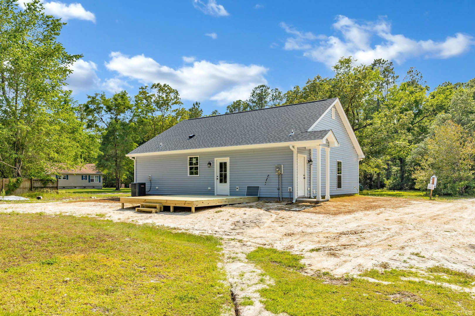 Wooden deck and white door with glass pane on blue house, dirt path leading to entrance, grassy yard, mature trees in background under partly cloudy sky