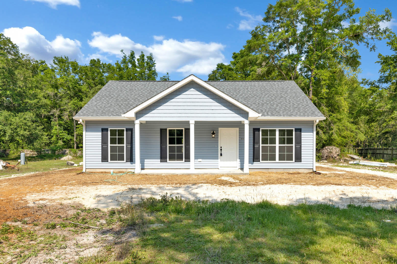 Two-story cottage-style home with white front door, black shutters, large windows, and mature trees in the background