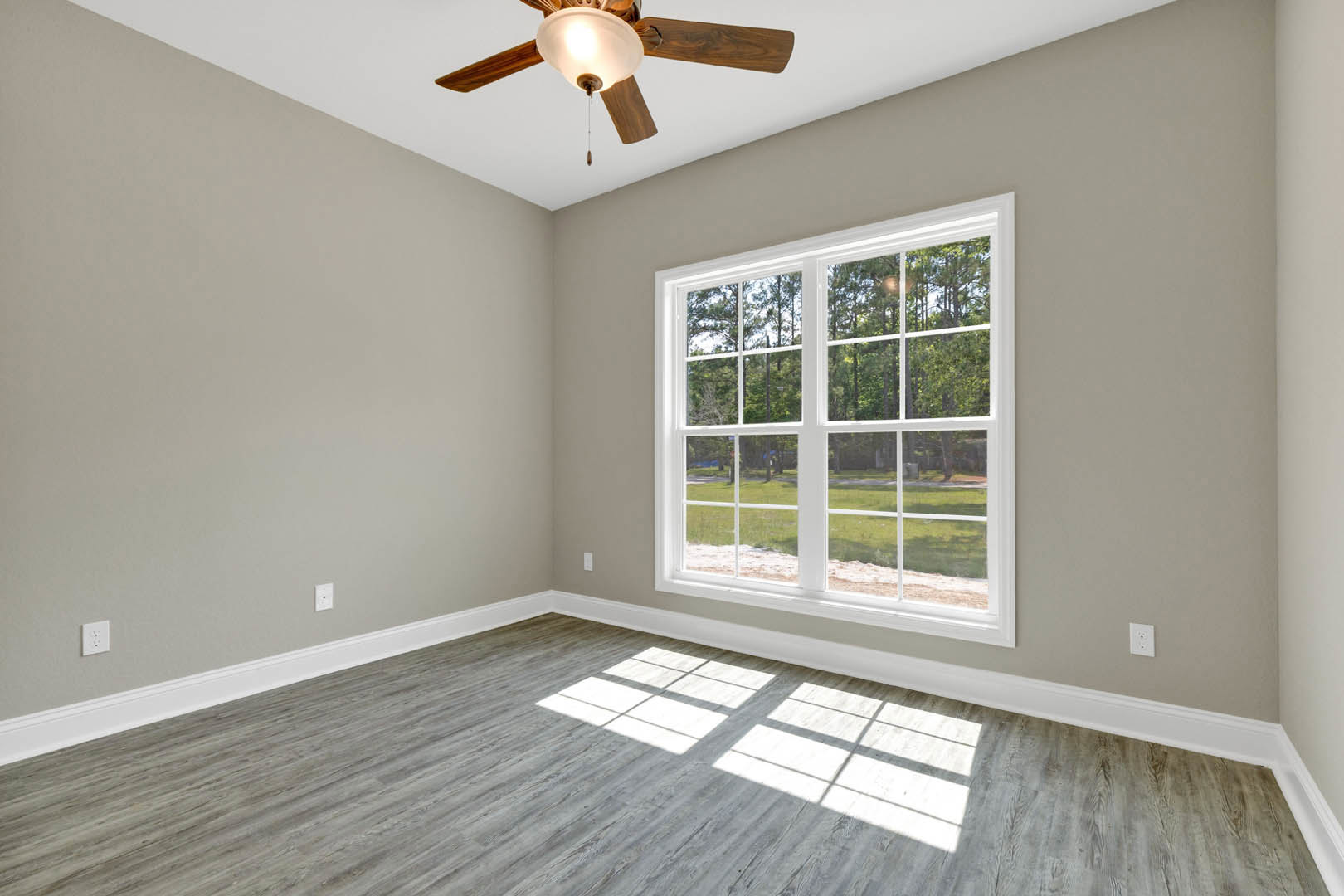 Wood floor room with white walls, ceiling fan with light fixture, large window letting in natural light, trees visible outside, crown molding along ceiling.