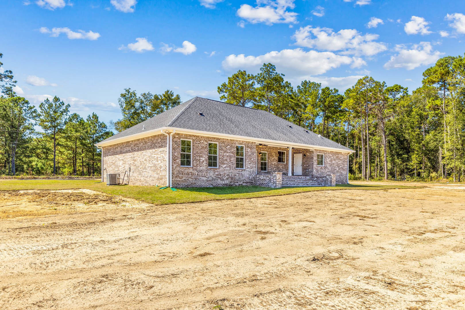 Red brick house with multiple windows, surrounded by a dirt road and grassy field, mature trees and blue sky with scattered clouds in the background
