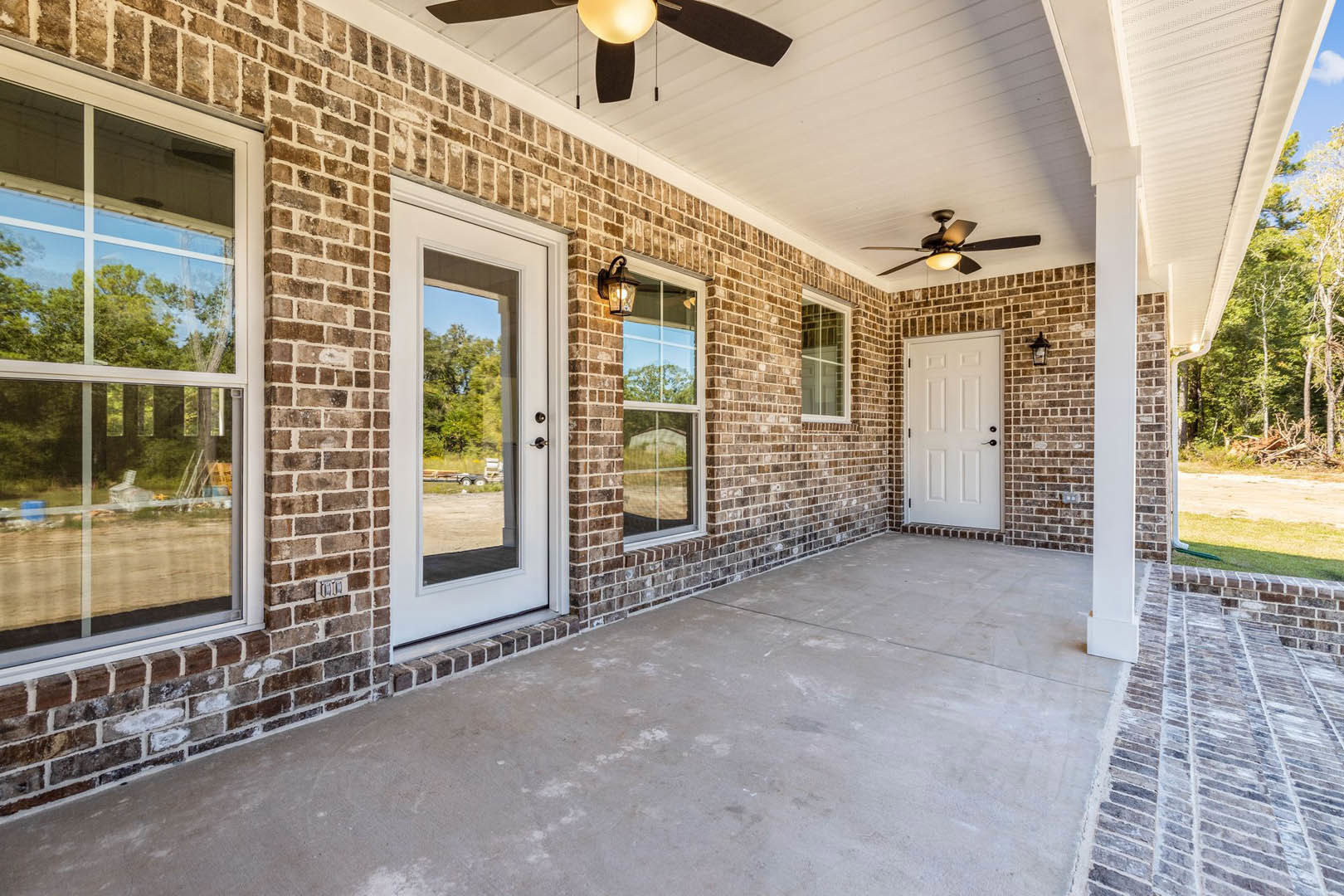 Red brick house exterior with white door featuring black handles, multiple glass-pane windows, ceiling fan on covered porch, and view of trees and trailer in background
