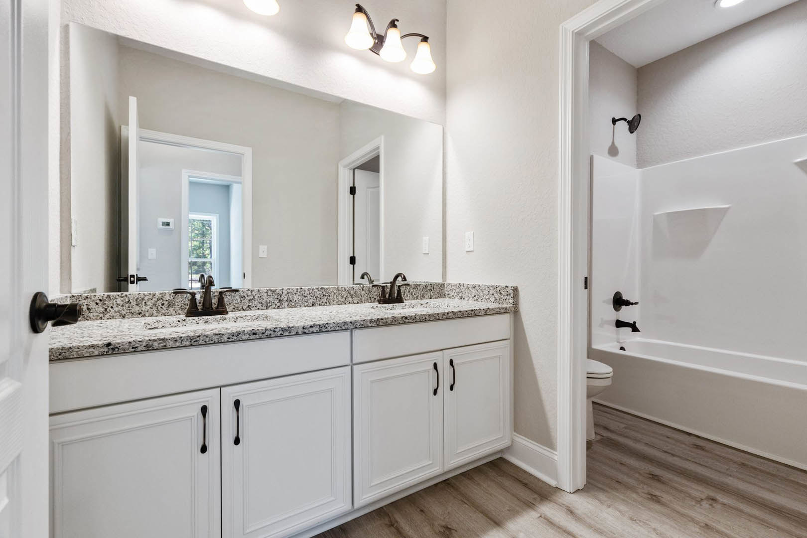 Marble countertop with undermount sink, white cabinetry featuring black handles, three-light fixture above mirror, white bathtub with wall-mounted shower head, toilet adjacent to