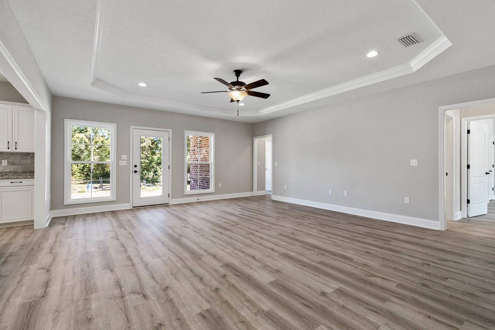 Ceiling fan with light fixture, hardwood floor, white door with black handles and window, large window showing trees outside, neutral walls and plaster ceiling