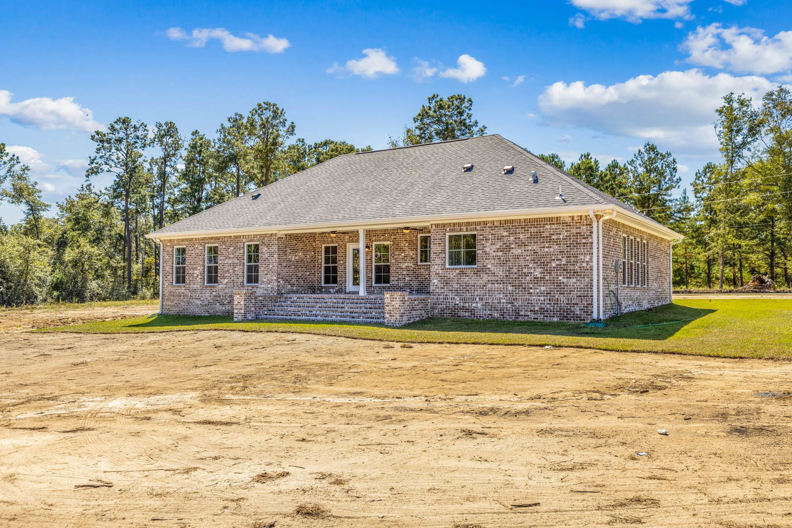Red brick house with covered porch, large windows, dirt yard with sparse grass, mature trees in background, cloudy sky overhead