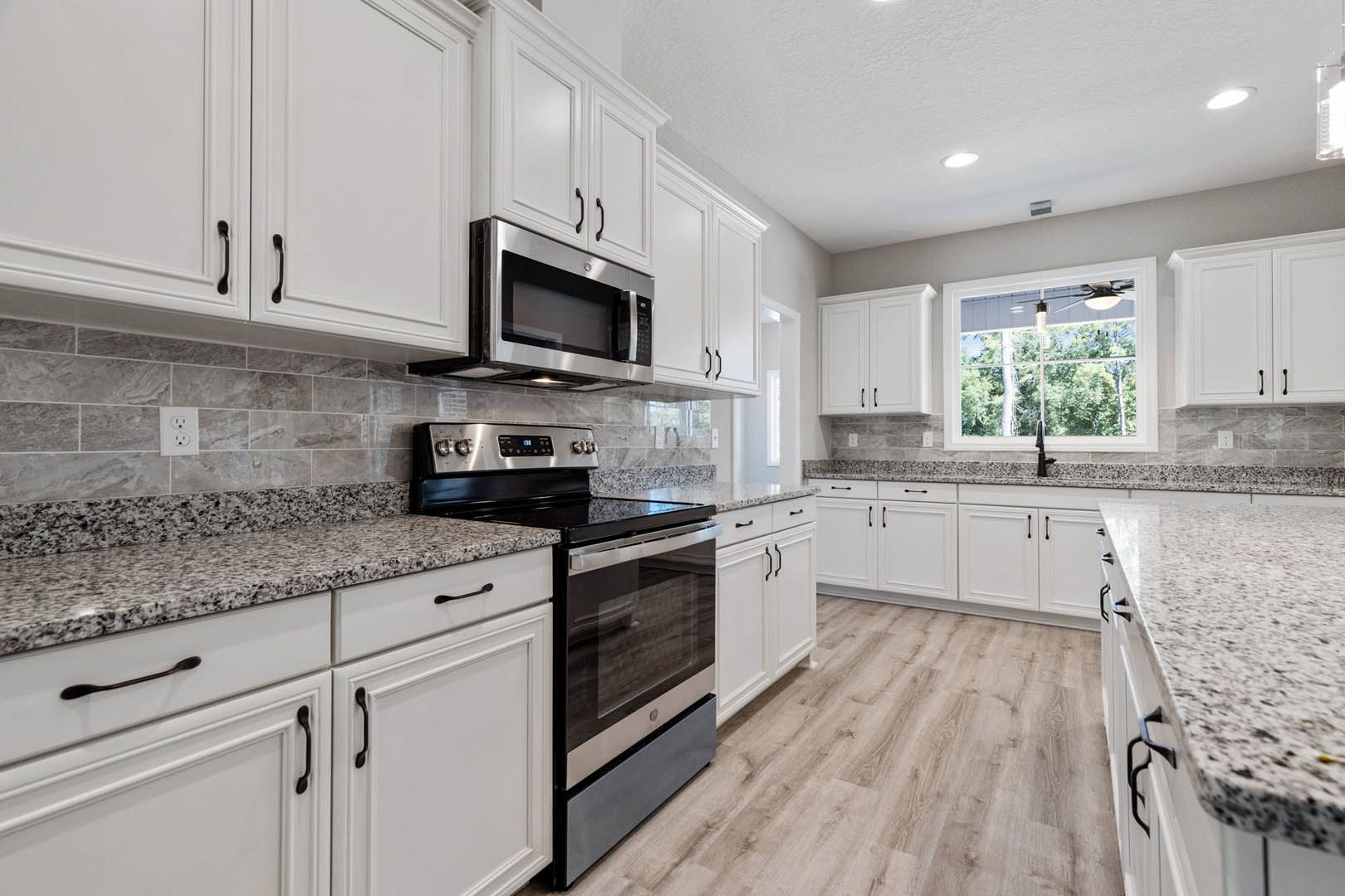 White kitchen cabinets with granite countertops, stainless steel stove and oven, black-door microwave, window above sink with fan and light bulb, white electrical outlet on