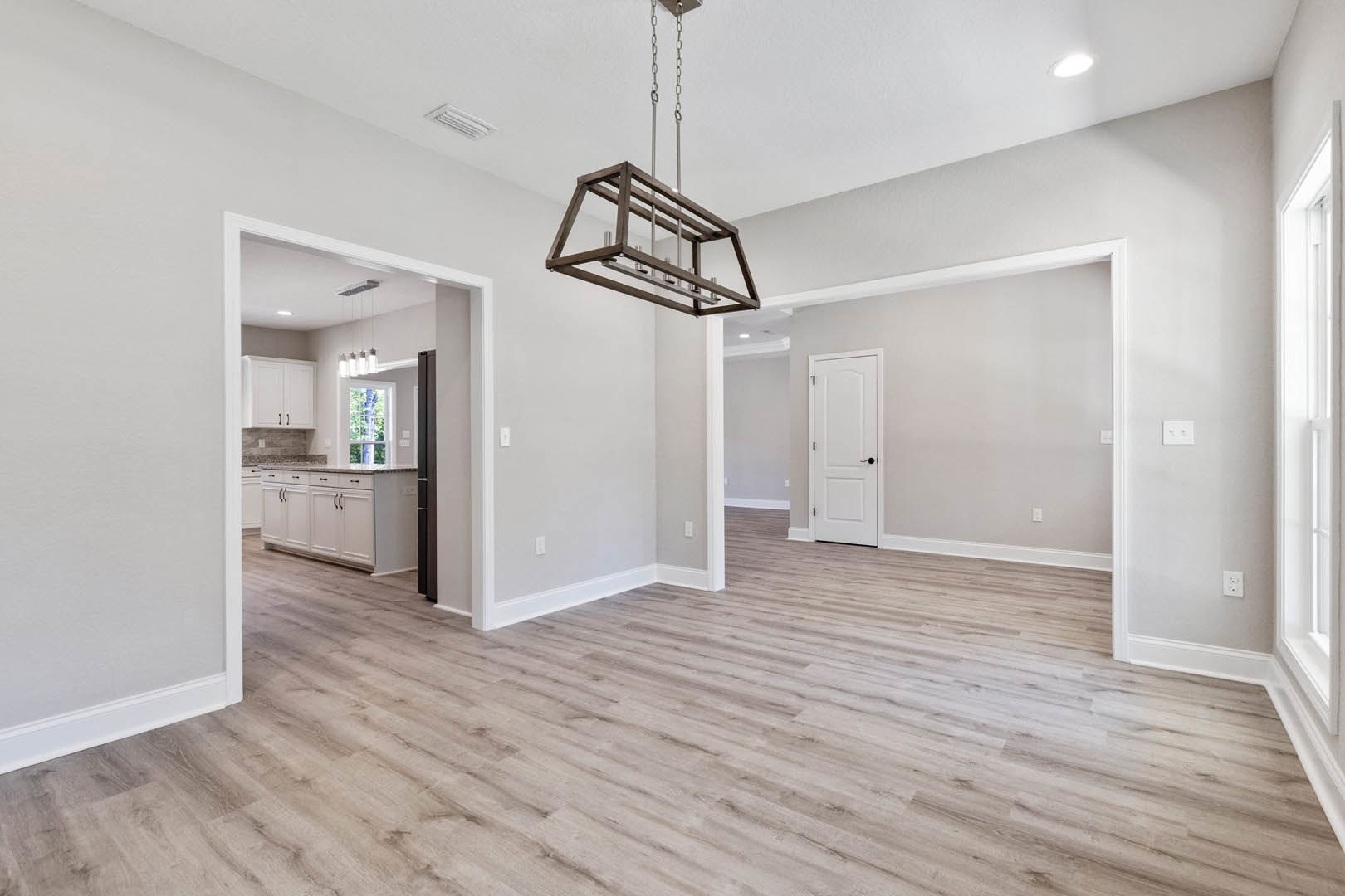 Open concept room with hardwood flooring, white trim, and white kitchen cabinets with black handles; wooden light fixture with metal rods and chain hangs from ceiling; white door