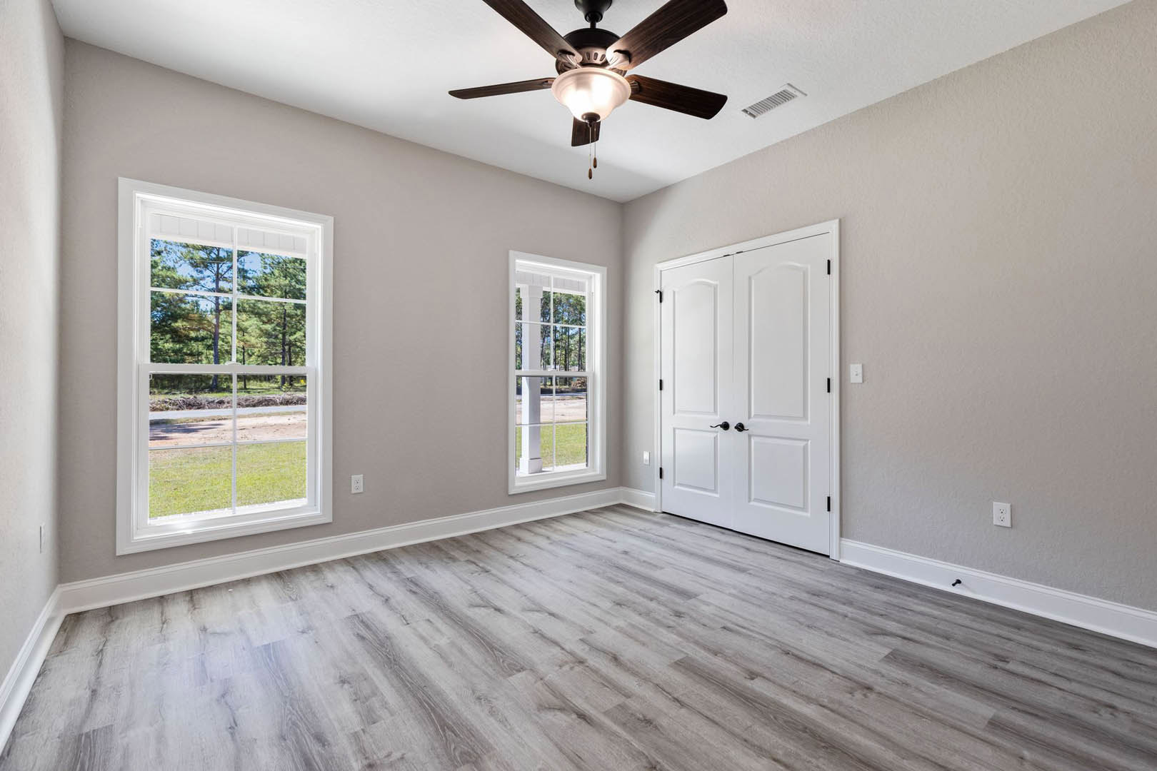 Ceiling fan with light fixture, two windows showing trees outside, white double doors with black handles, wood laminate flooring, white walls