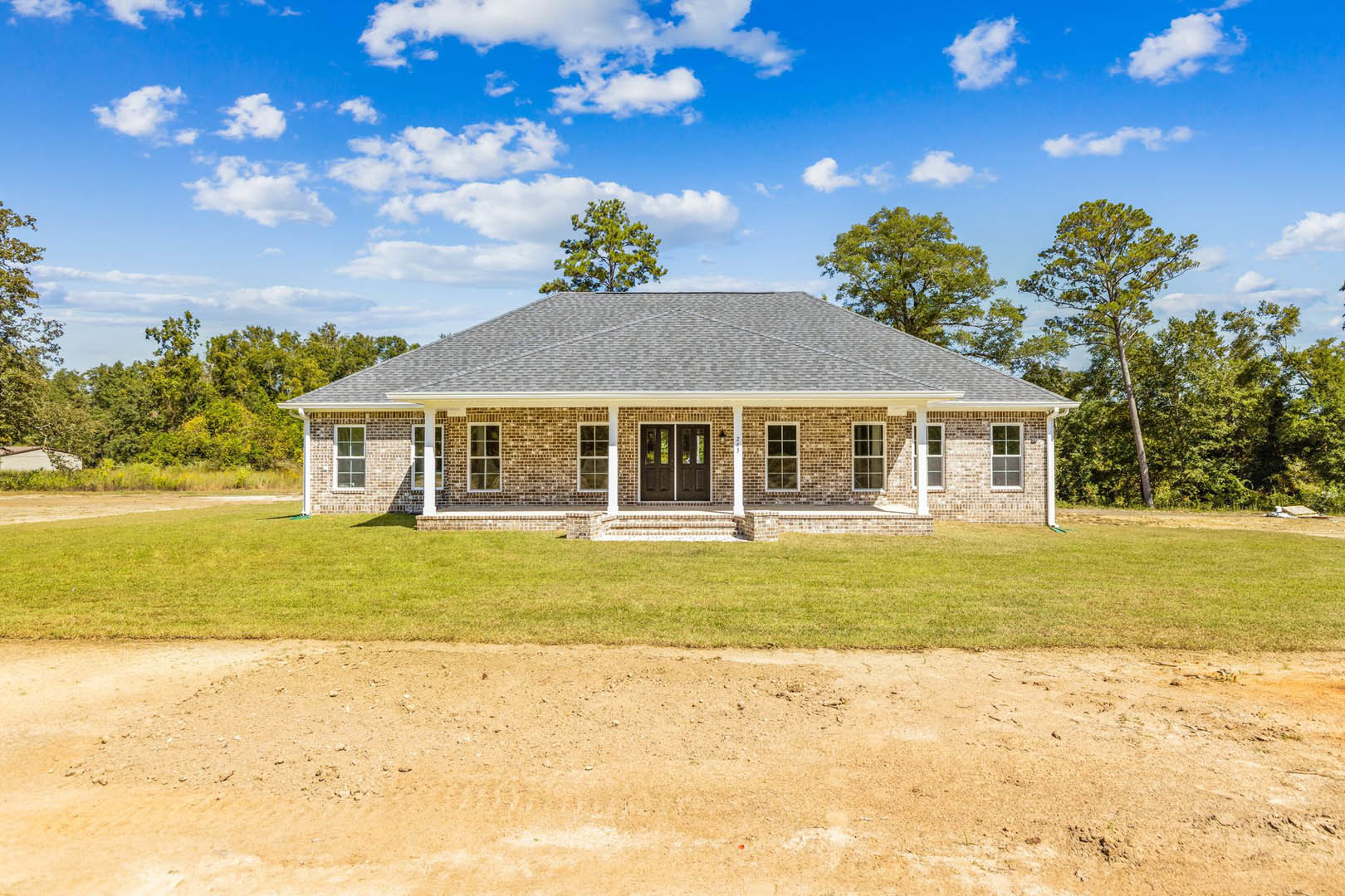 Front view of a brick house with double doors, dirt road leading to the entrance, grassy lawn, and mature trees in the background under a cloudy sky