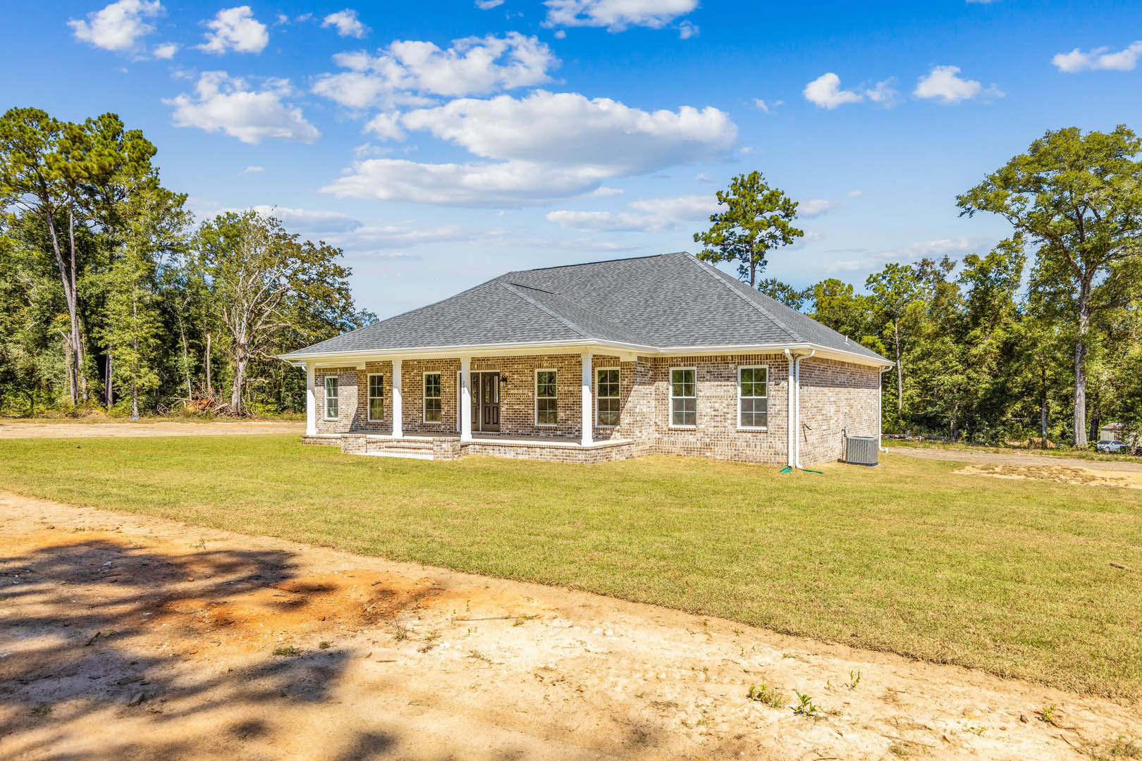 Red brick house with white-framed windows, expansive green lawn, white fence, dirt path along grass, leafy tree, and grey air conditioner unit beside exterior wall