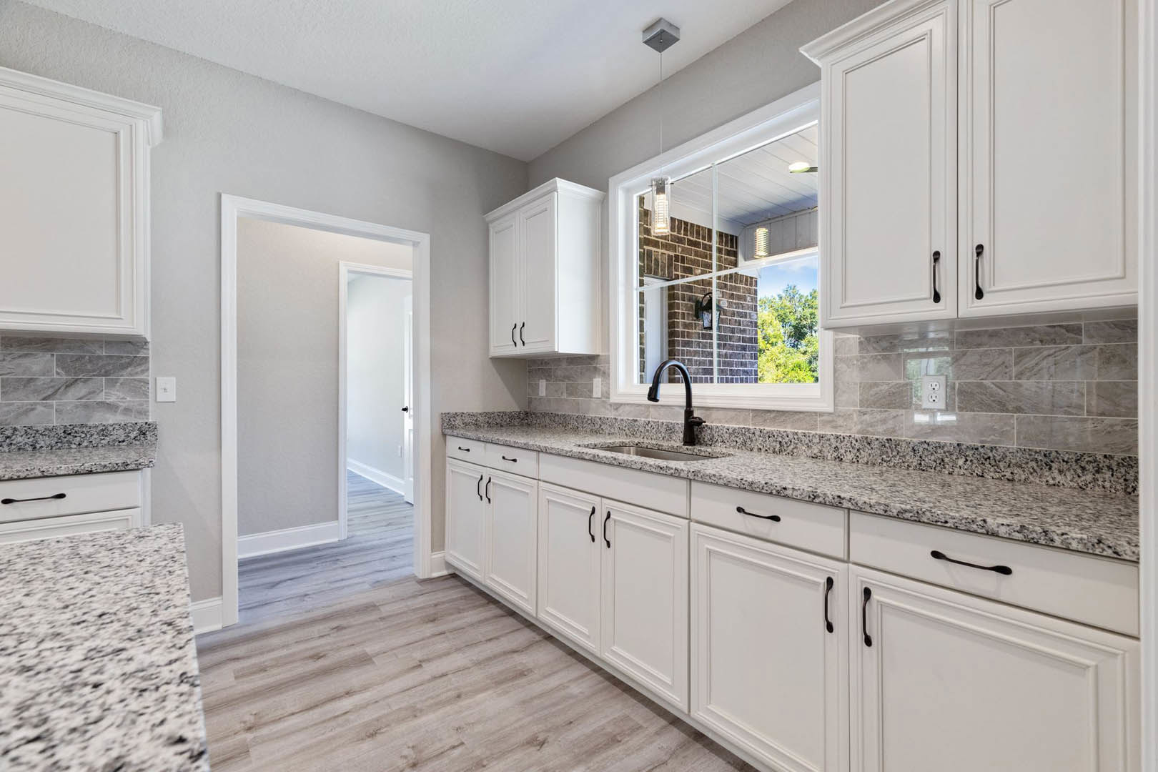 Kitchen with white cabinets, granite countertops, stainless steel sink, black cabinet handles, white tile flooring, window with brick wall view, modern light fixture
