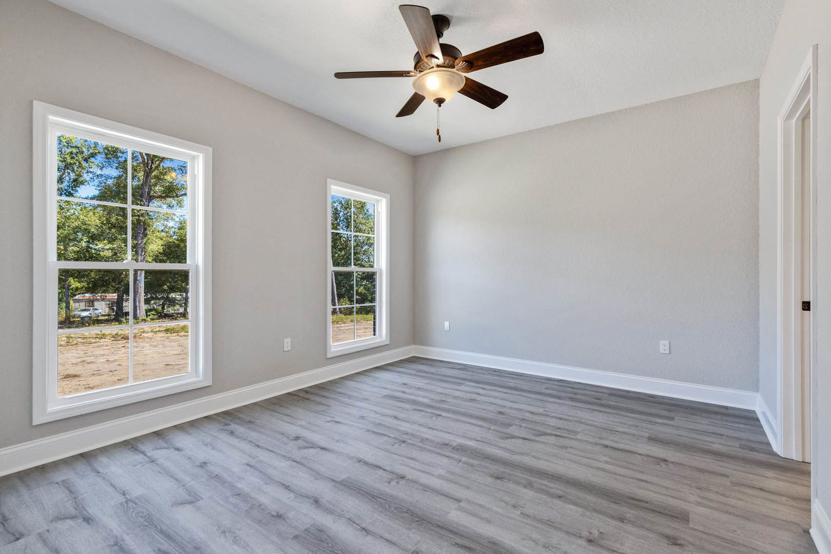 Ceiling fan with light fixture, grey wood laminate flooring, white baseboard trim, large windows showing trees and parked car outside