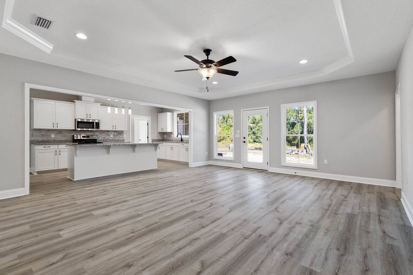 Spacious room with hardwood flooring, ceiling fan with light fixture, white door with glass window, large window overlooking a tree, built-in cabinetry, and stainless steel oven