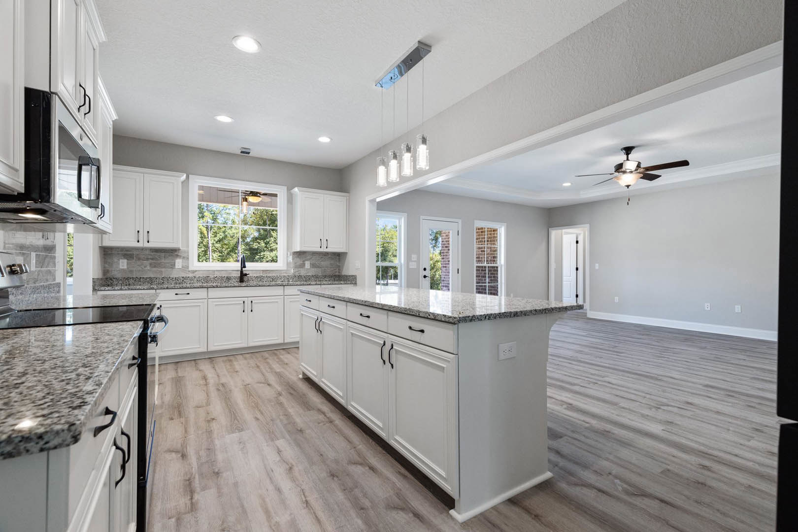 Spacious kitchen featuring a large central island with white cabinetry, wood flooring, stainless steel sink, and a ceiling fan with light; windows reveal trees outside, and white