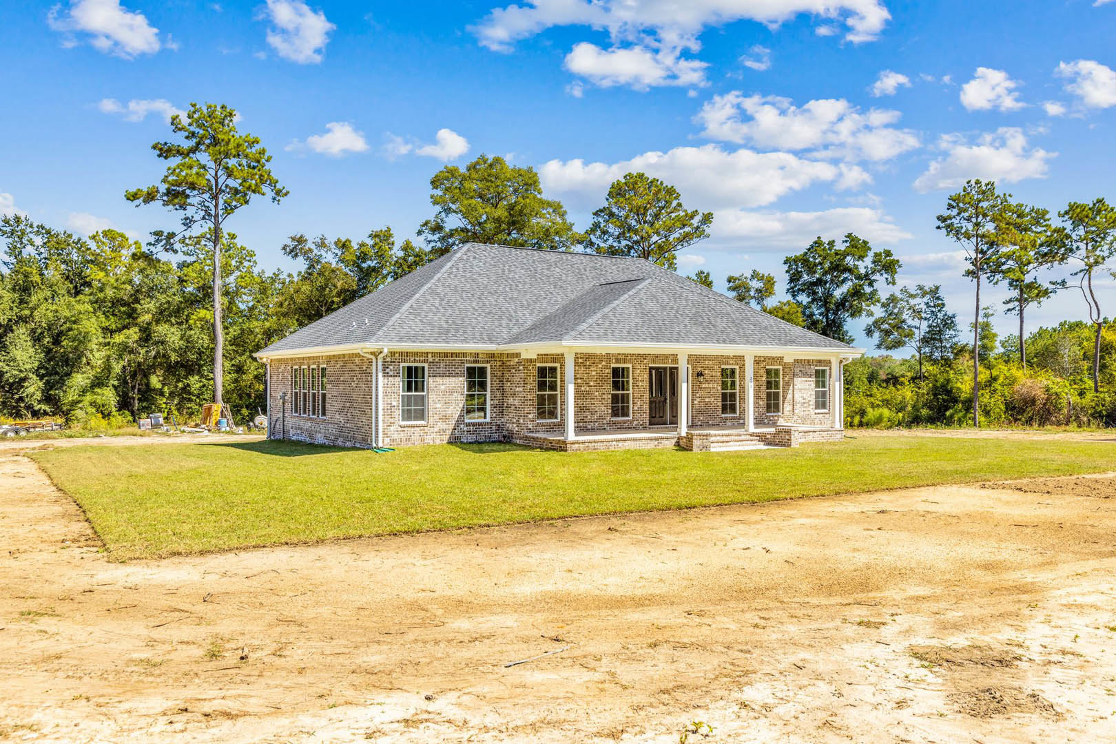 Brick house with white columns, surrounded by tall green trees, dirt road bordered by grass leading to the entrance, cloudy sky overhead