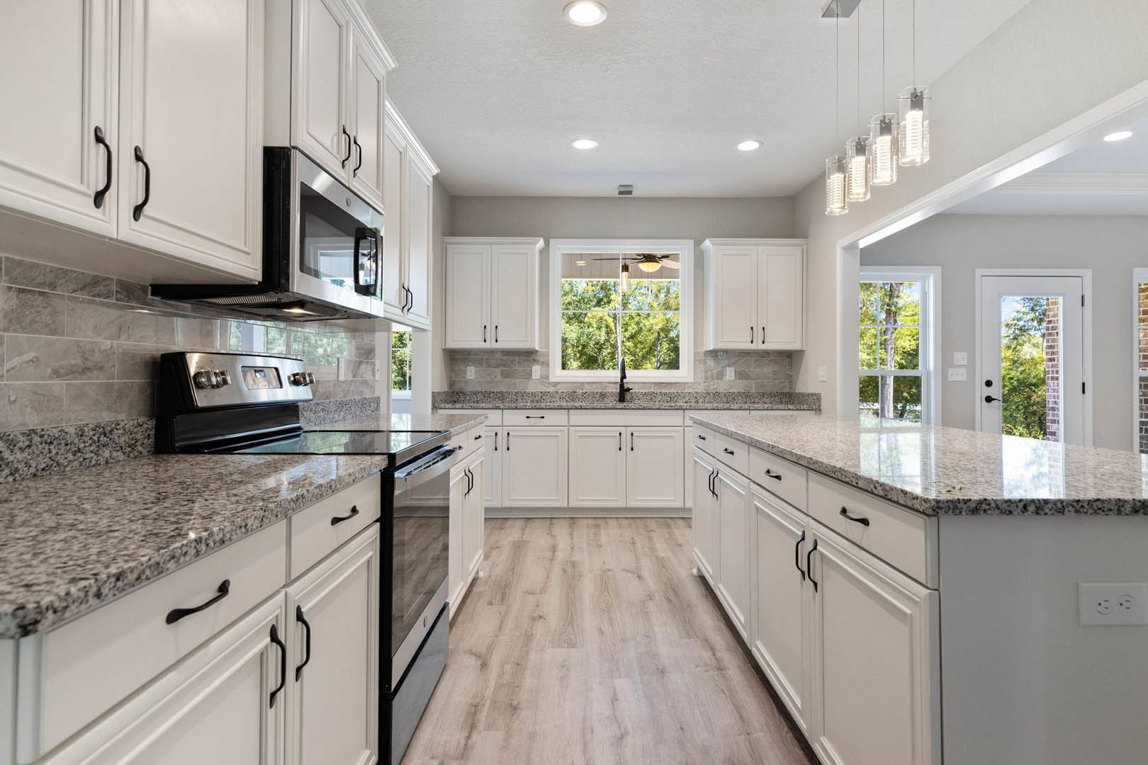 White kitchen cabinets and granite countertops, stainless steel microwave, stove with visible knob, wood flooring, white door with glass window, window above sink showing fan and
