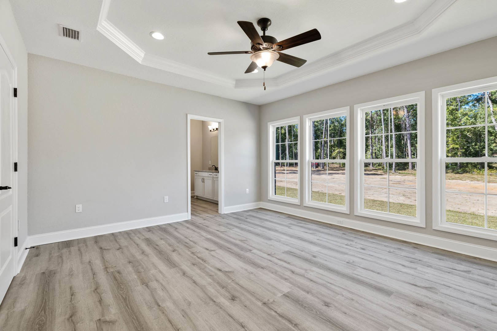 Spacious room with wood flooring, ceiling fan with light, multiple large windows framed by white molding, trees visible outside