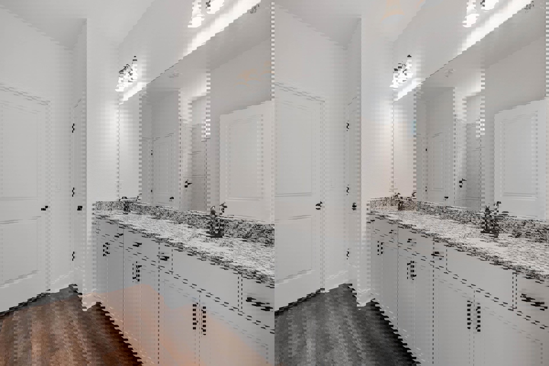Bathroom with white cabinets, expansive mirror above countertop, wood floor with white trim, white doors featuring black hardware, and three-light fixture mounted above the mirror