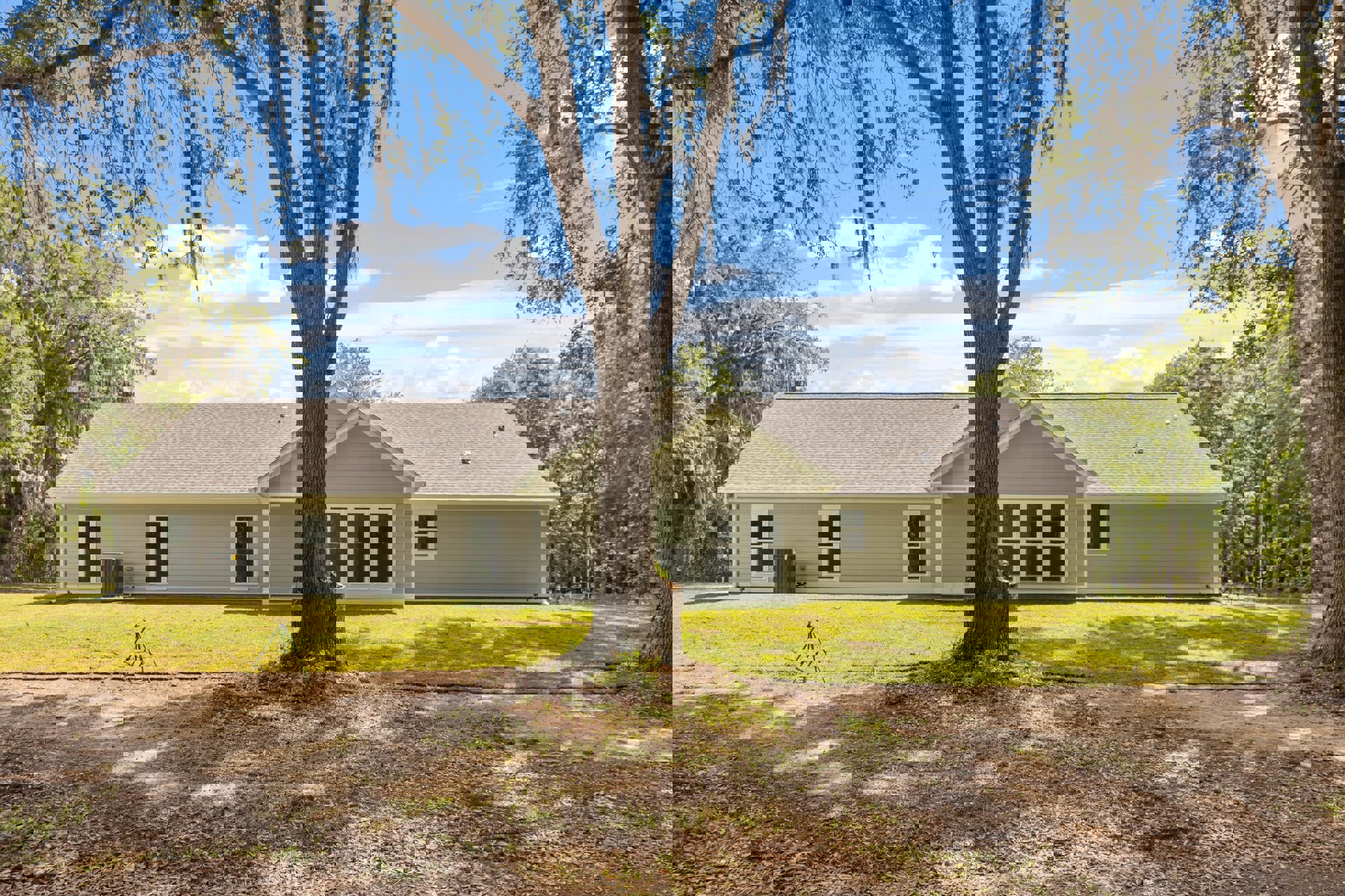 Two-story house with white-framed windows, air conditioner unit beside exterior wall, large tree trunk in grassy yard, cloudy sky overhead
