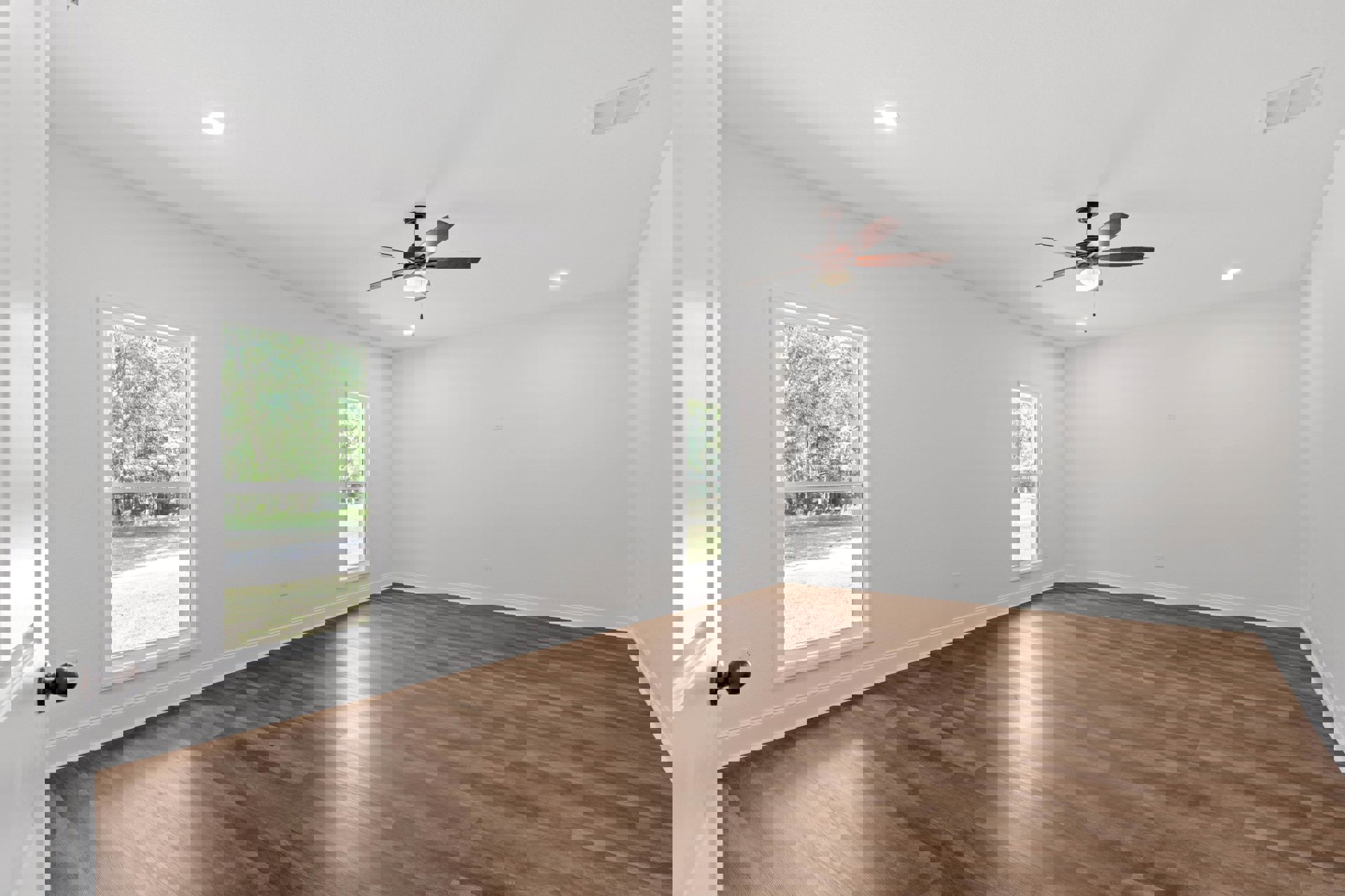 Ceiling fan with light fixture, wood laminate flooring, white plaster walls, large windows showing trees and yard, ceiling vent