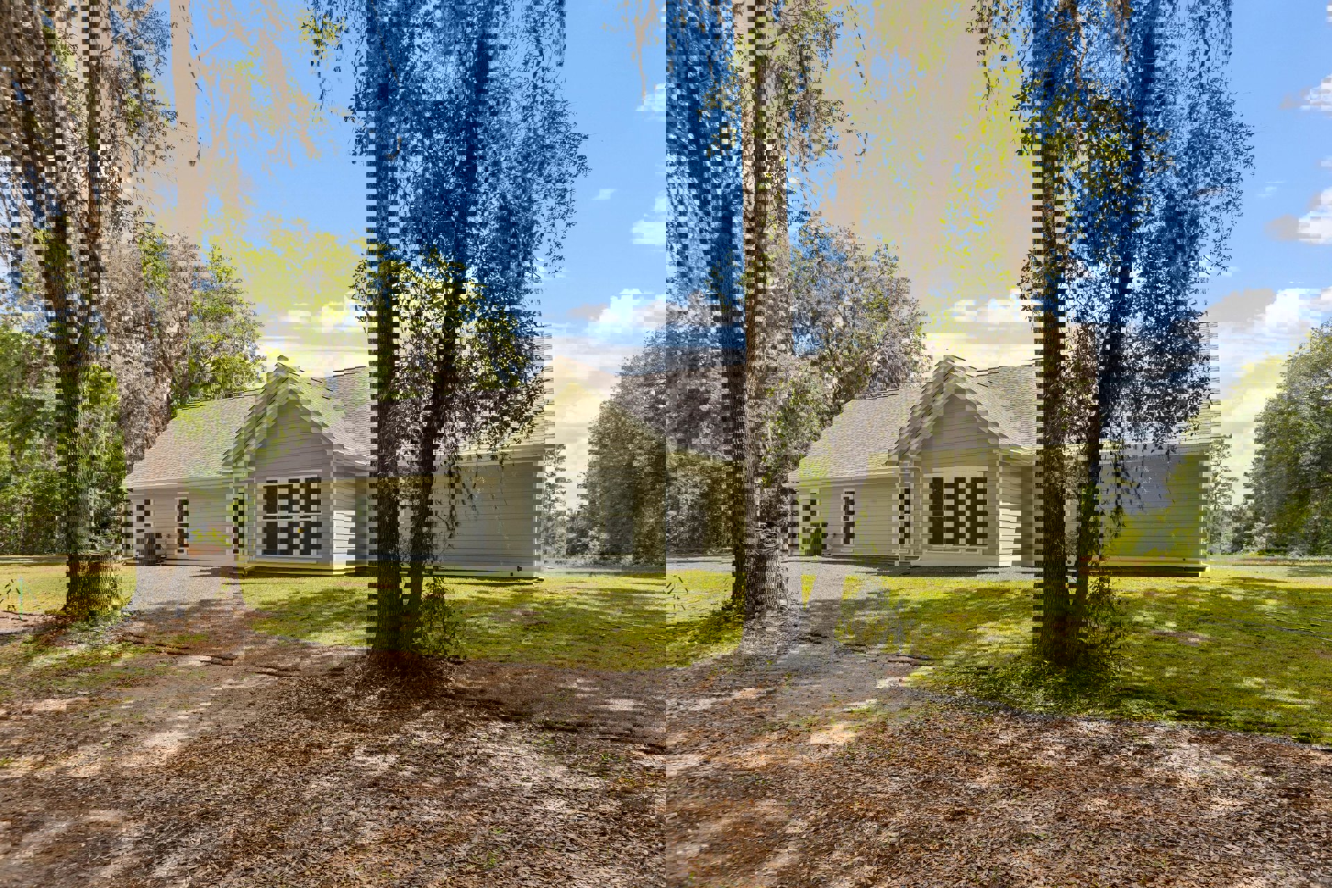 White farmhouse with large windows, green lawn, mature trees behind and in front, moss-covered tree trunk, cloudy sky overhead