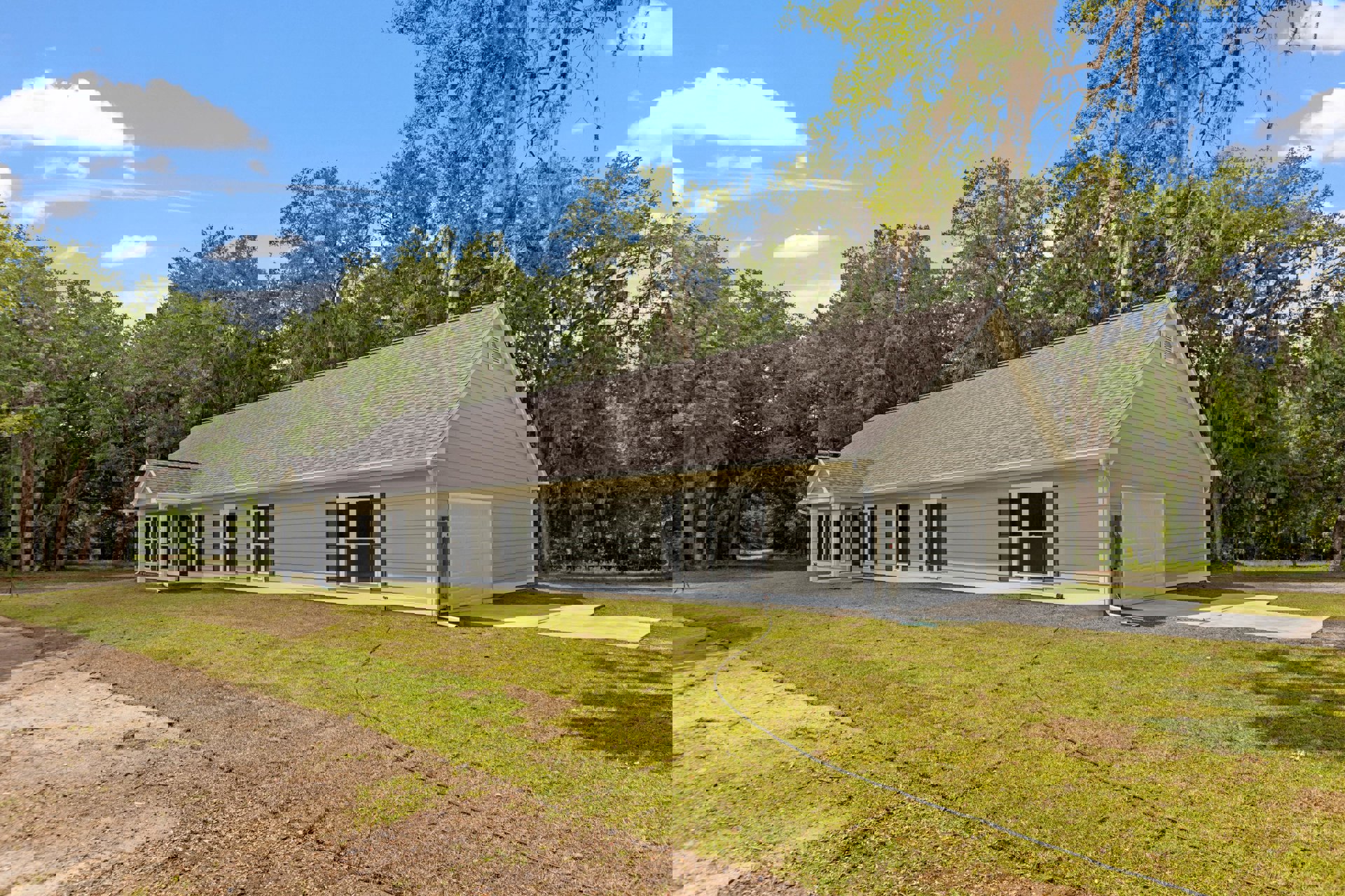 Two-story house with attached garage, white siding, asphalt shingle roof, green grass lawn, garden hose on the yard, mature trees in the background, partly cloudy sky