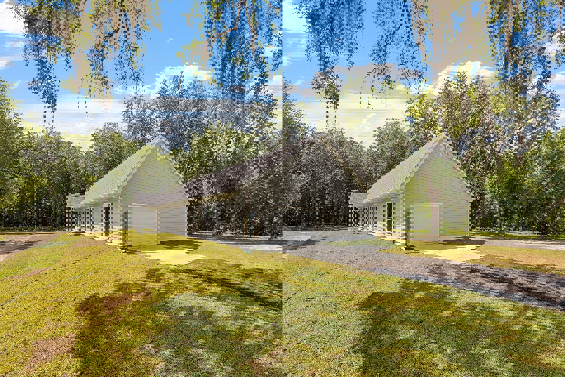 White farmhouse-style home with attached two-car garage, gray shingle roof, large windows, and manicured green grass lawn bordered by mature trees under partly cloudy sky