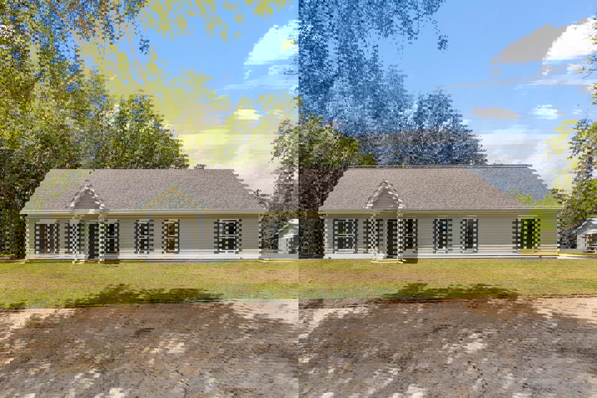 Two-story house with white-framed windows, black front door, green lawn, mature trees casting shadows, and partly cloudy sky