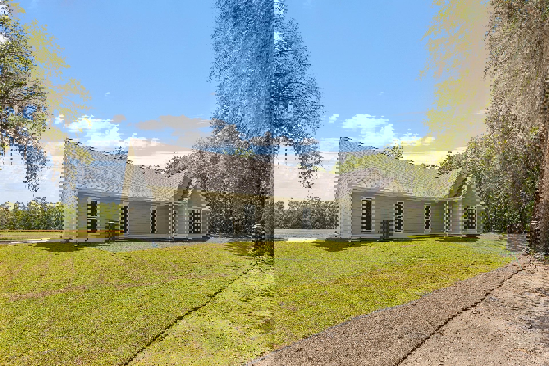 Two-story house with white-framed windows, gray siding, heat pump unit beside exterior wall, green lawn with garden hose, mature trees with moss, cloudy sky overhead