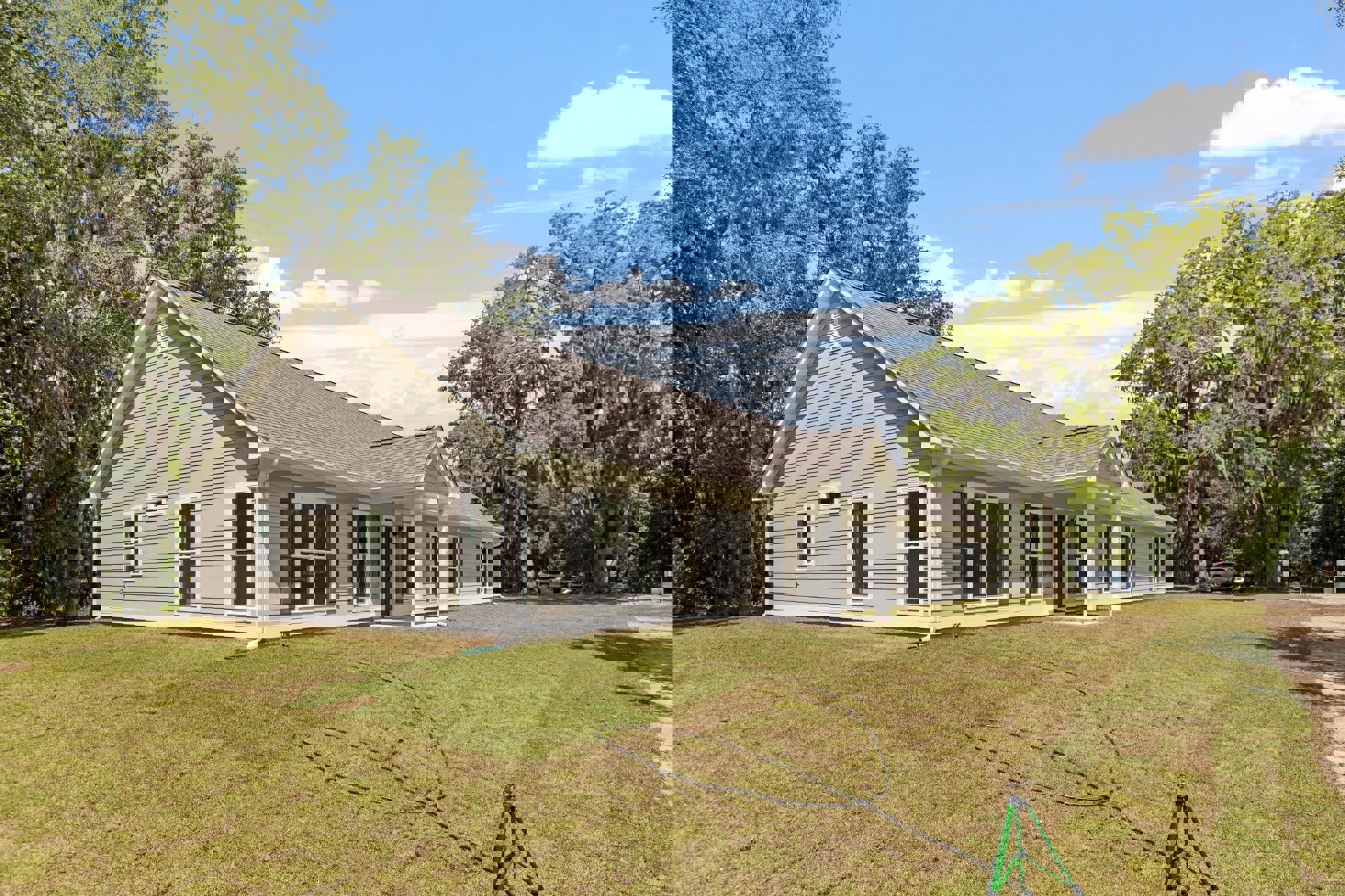 Two-story farmhouse with white siding, large windows, manicured lawn, mature trees, garden hose on grass, and a car parked in the driveway