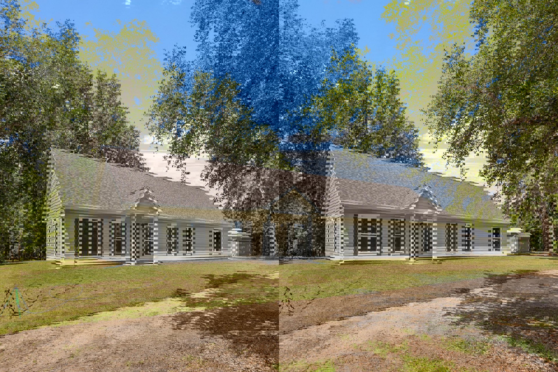 Two-story house with white-framed windows, covered front porch, dirt path leading to entrance, surrounded by grass and mature trees under partly cloudy sky