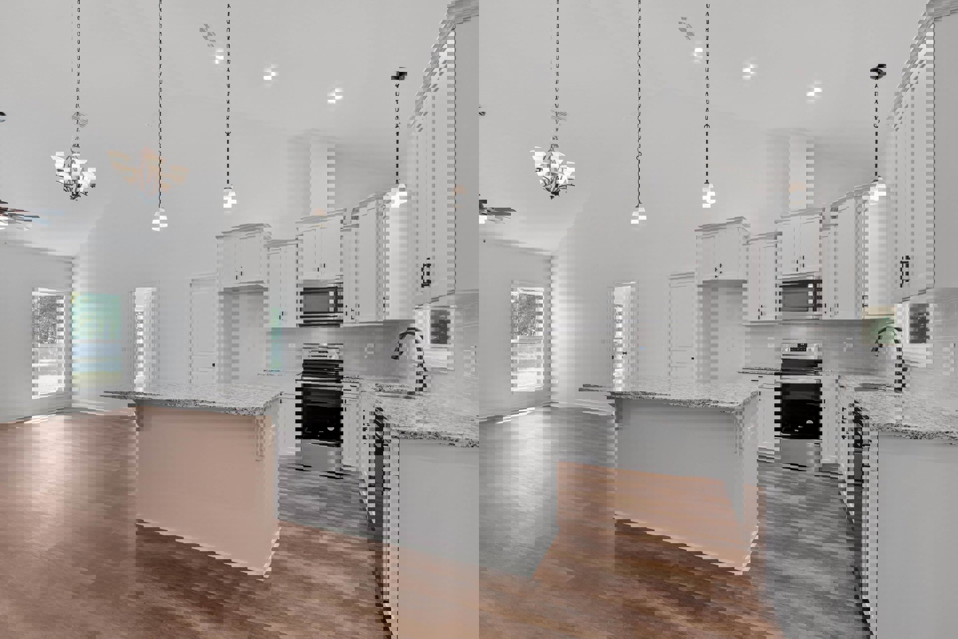 Spacious kitchen featuring a large marble-topped island, wood flooring, white cabinetry, stainless steel stove, open microwave, modern chandelier, and a window overlooking trees