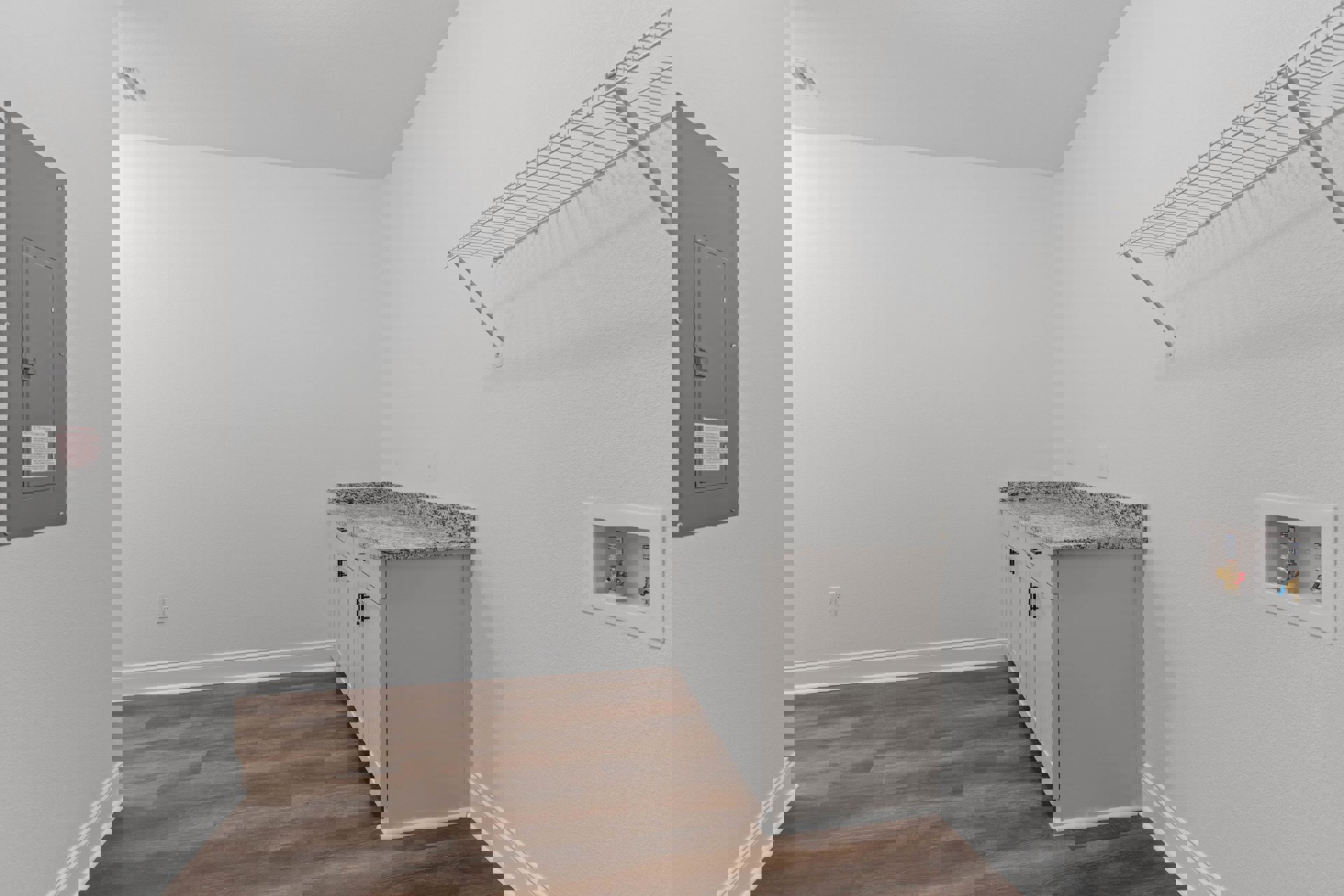 Kitchen with granite island countertop, wood flooring, white plaster walls, white wire rack mounted on wall, grey panel with square opening, and notice board displaying red text.